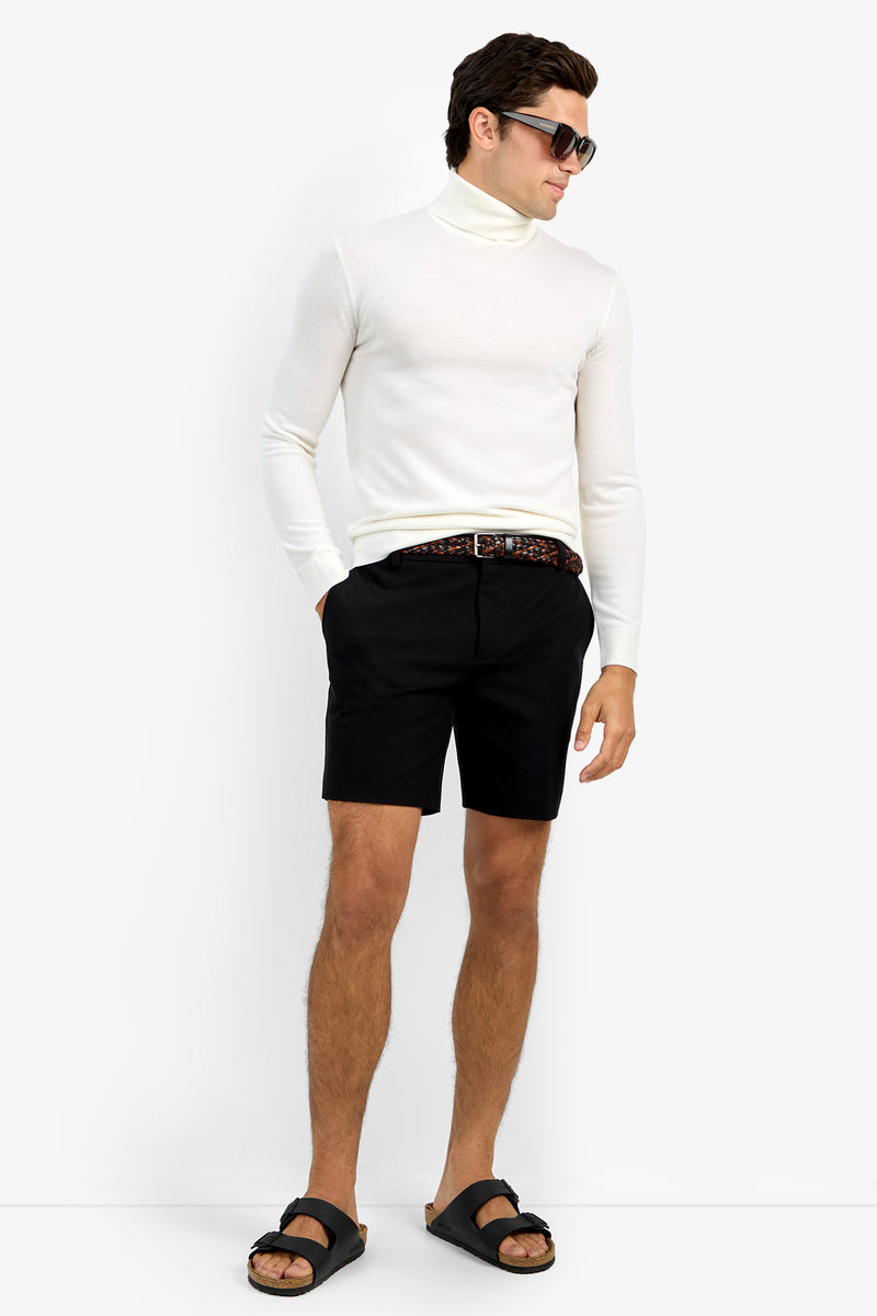 A man stands against a plain white background wearing the Roy Long Sleeve Turtleneck, black shorts with a patterned belt, black sandals, and sunglasses, hands in his pockets.
