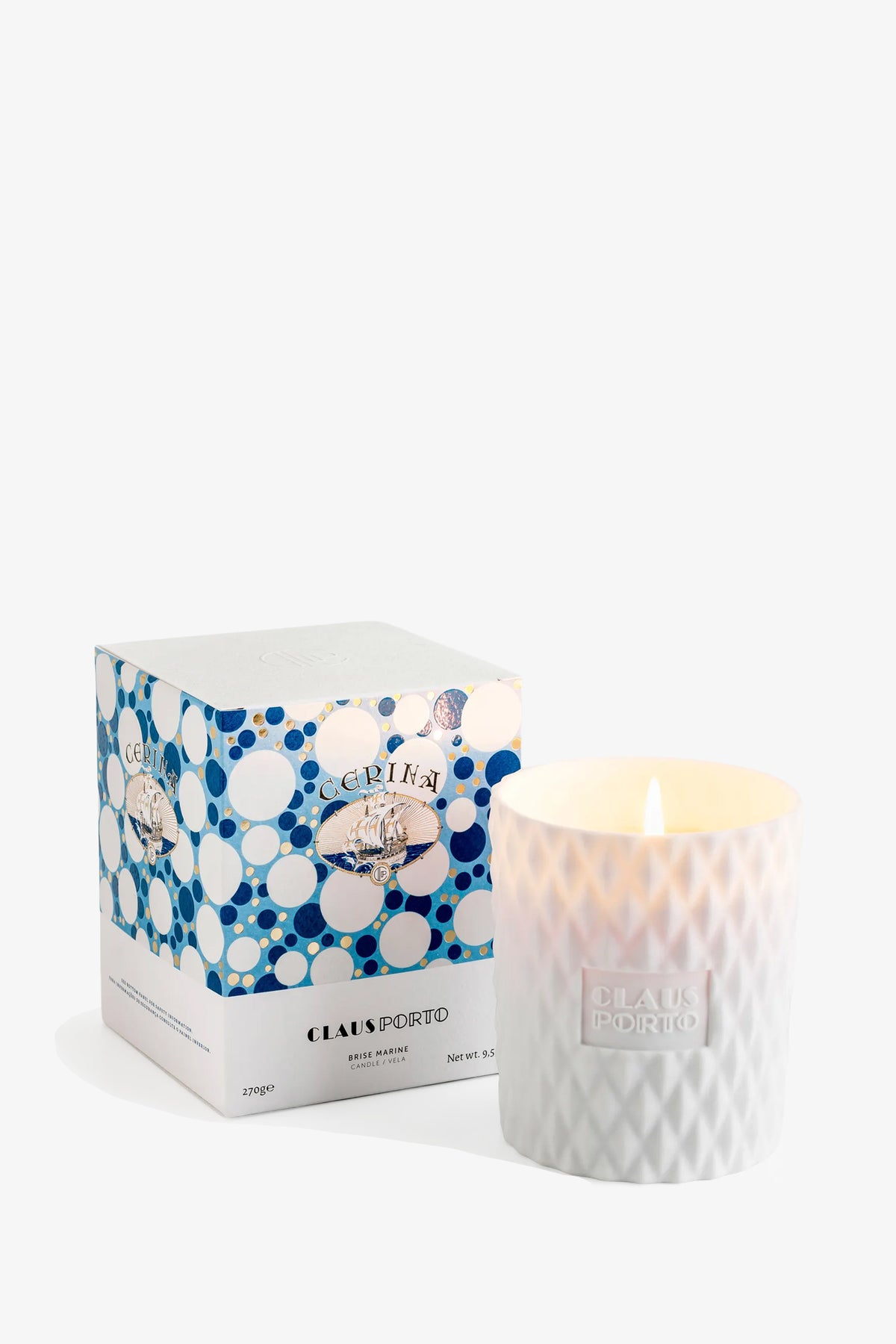 A lit Claus Porto Cerina Candle in a textured glass holder sits next to its blue-and-white patterned box.