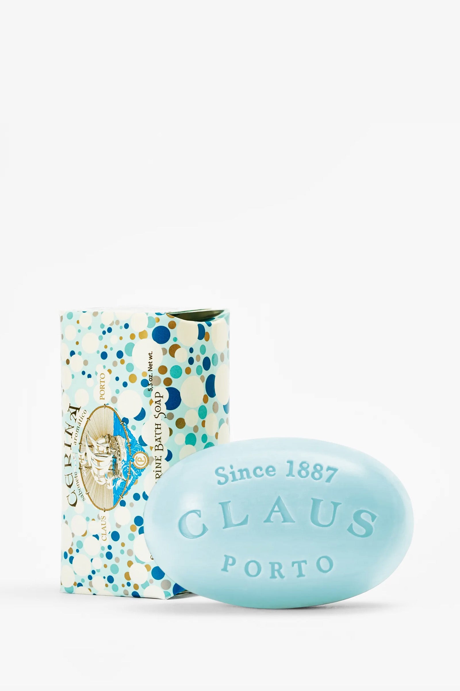 A light blue, oval-shaped Claus Porto Cerina Soap bar labeled “CLAUS PORTO Since 1887” is placed in front of a colorful, patterned soap box featuring blue, gold, and white dots.