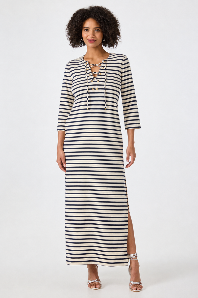 A woman with curly hair stands against a plain white background wearing the Catamaran Dress, a long-sleeve, ankle-length black and white striped dress featuring a lace-up neckline and side slit. She completes her look with silver strappy sandals.