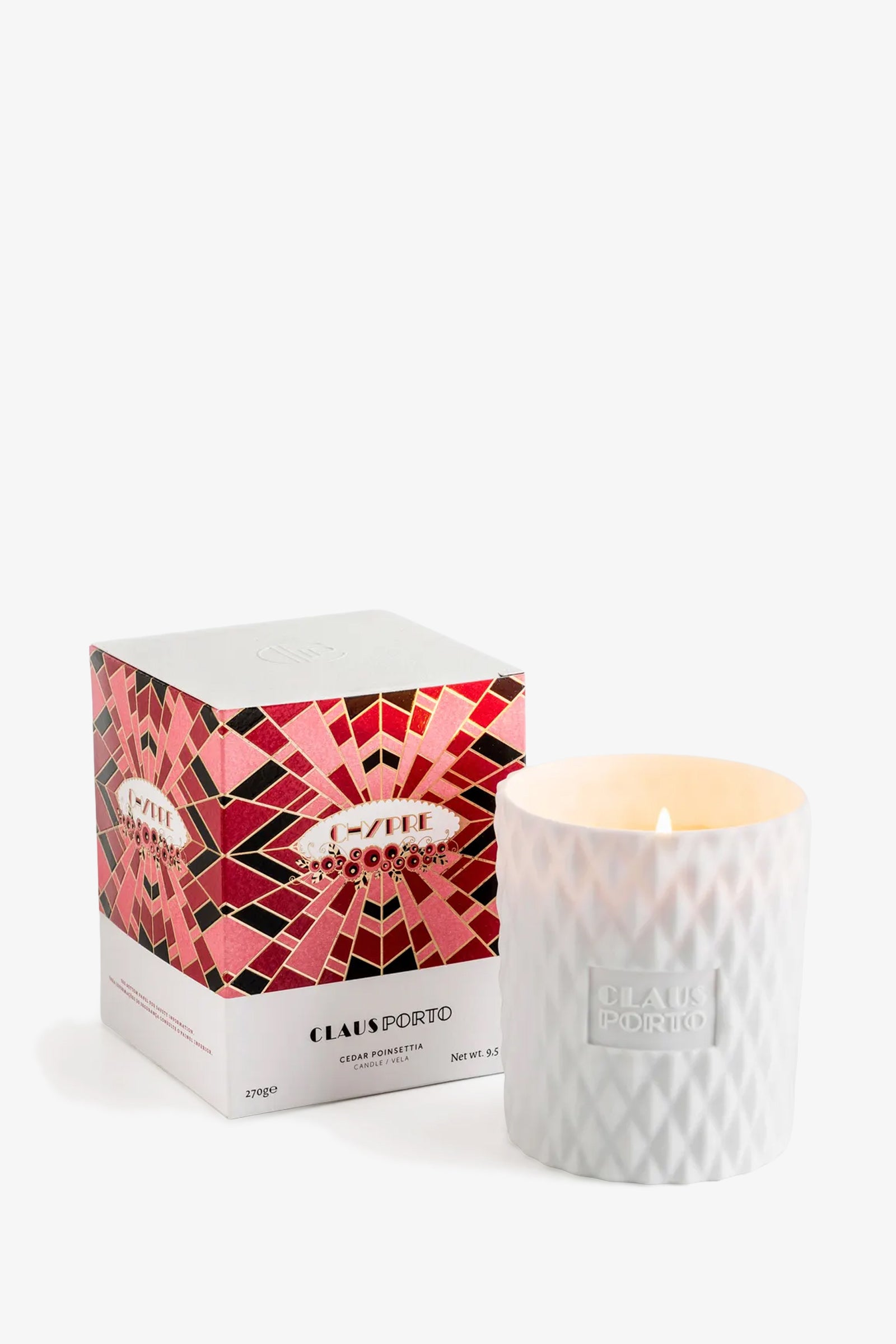 A lit Claus Porto Chypre Candle with a white textured finish in a matching holder sits next to its colorful, geometric-patterned box.