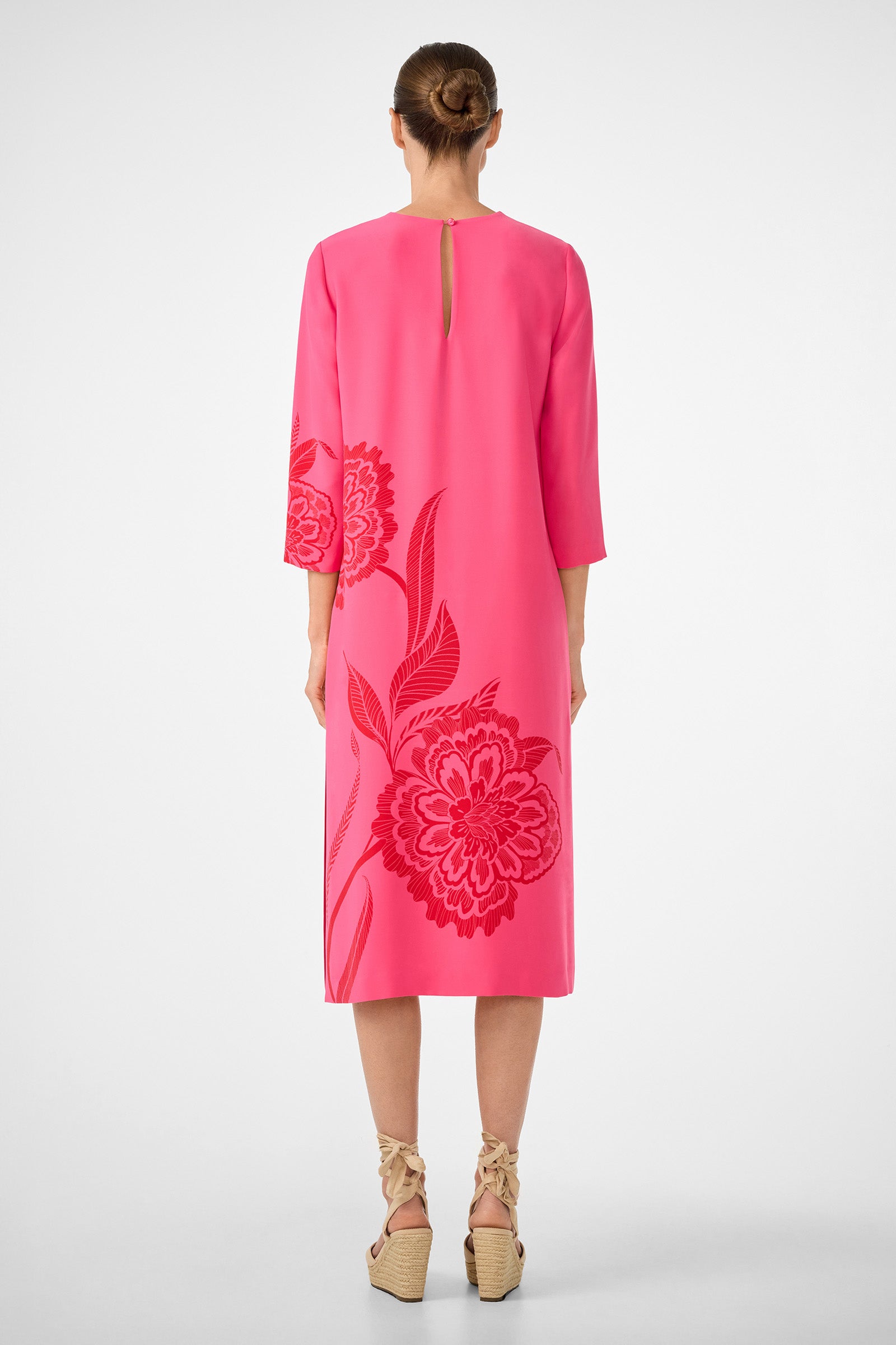 A woman faces away, wearing the Cointreau 2 Dress—a bright pink midi dress with floral patterns, elbow-length sleeves, and a small back keyhole detail. Her hair is in a bun, and she pairs the look with beige wedge sandals featuring ankle ties.