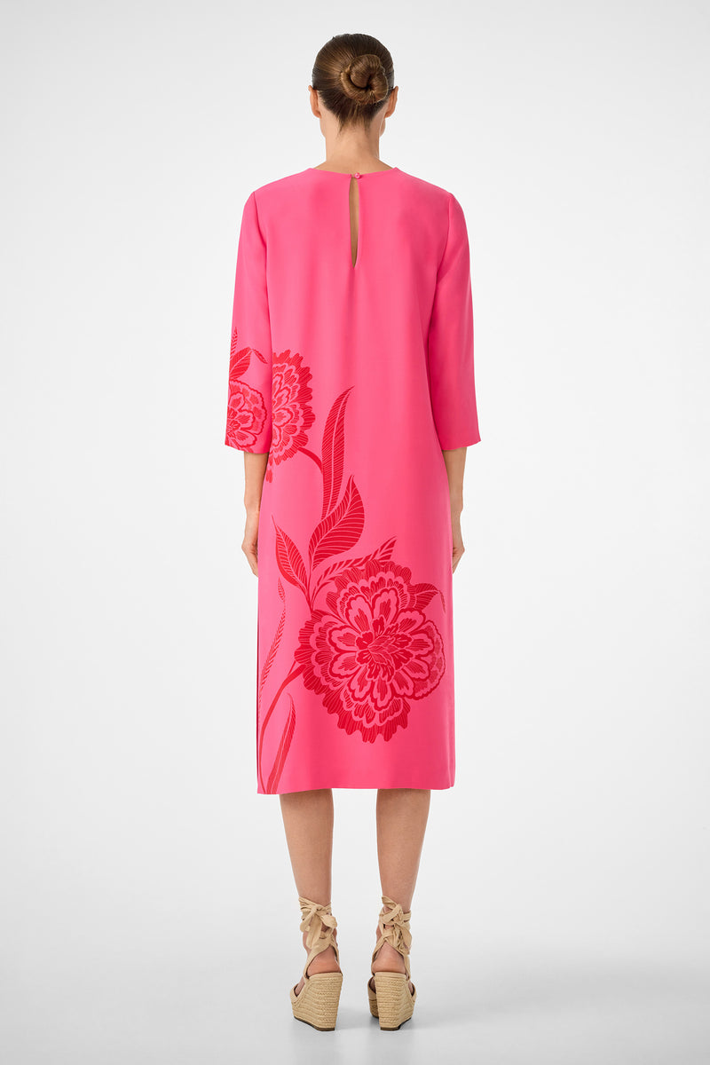 A woman faces away, wearing the Cointreau 2 Dress—a bright pink midi dress with floral patterns, elbow-length sleeves, and a small back keyhole detail. Her hair is in a bun, and she pairs the look with beige wedge sandals featuring ankle ties.