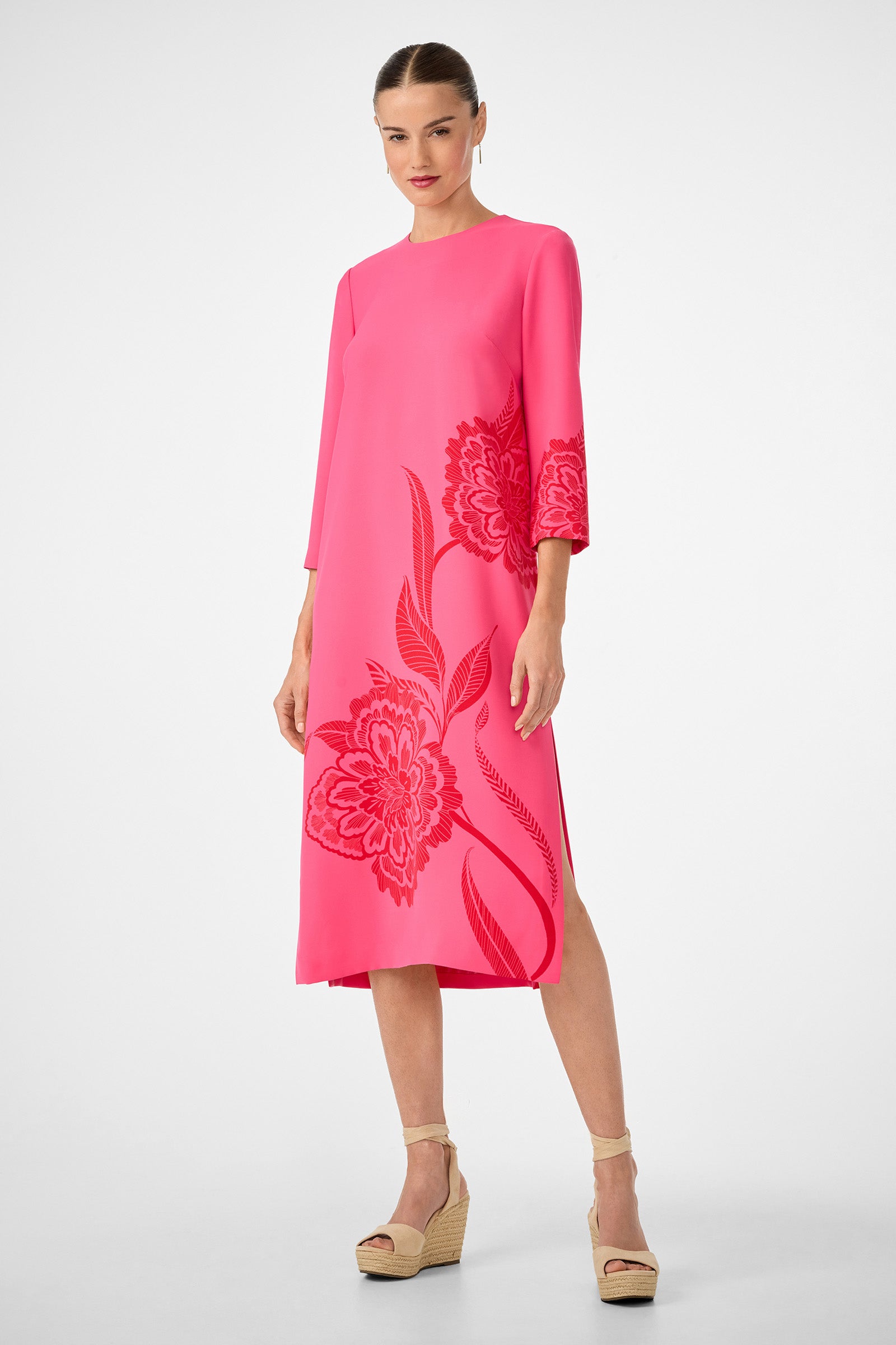 A woman wearing the Cointreau 2 Dress, a bright pink, mid-length dress with large red floral patterns and three-quarter sleeves, pairs it with beige wedge sandals against a plain white background.
