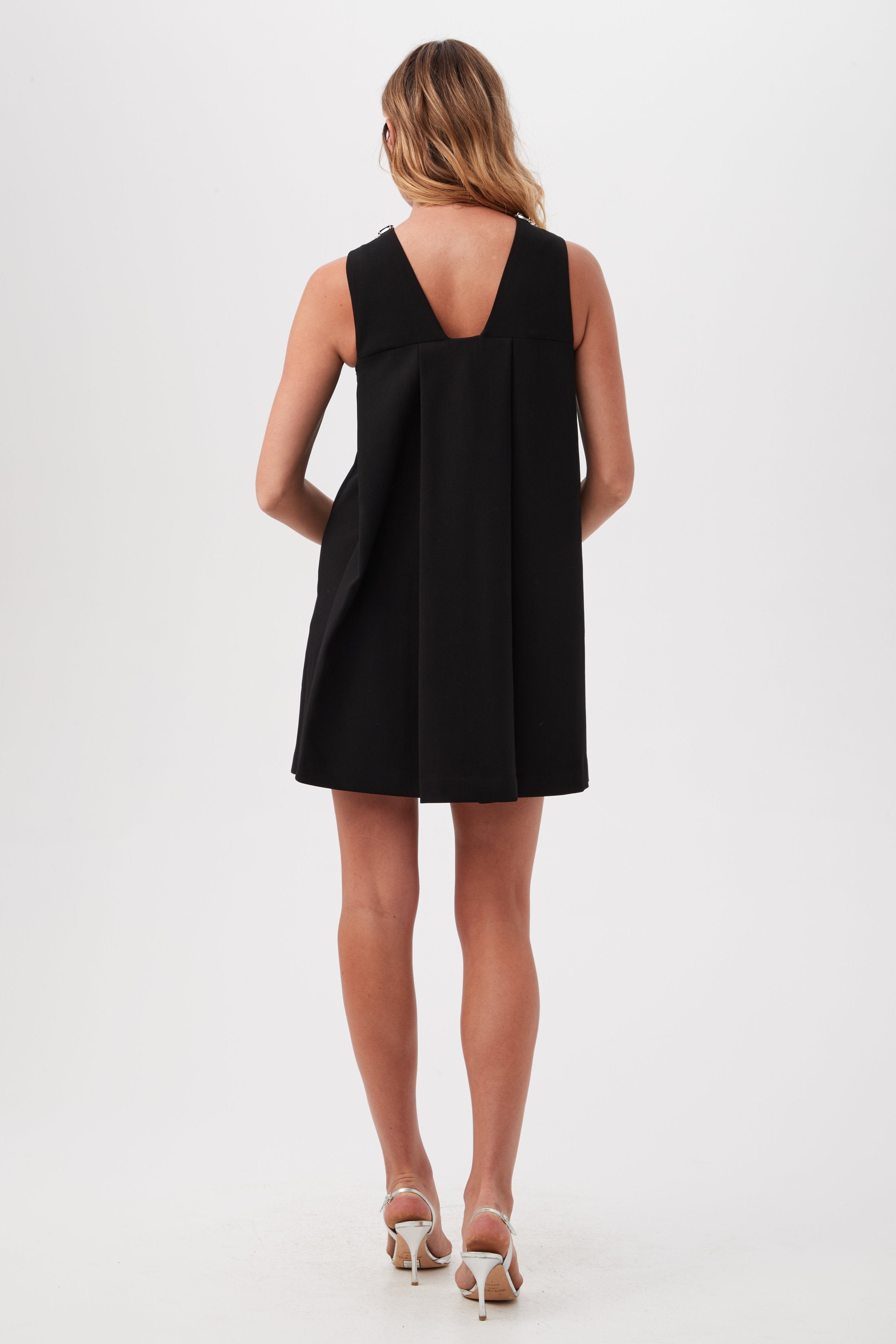 A woman with light brown wavy hair faces away, wearing the Beauty 2 Black Short Sleeveless A-Line Dress featuring a V-shaped back and paired with white high-heeled sandals, posed against a plain white background.