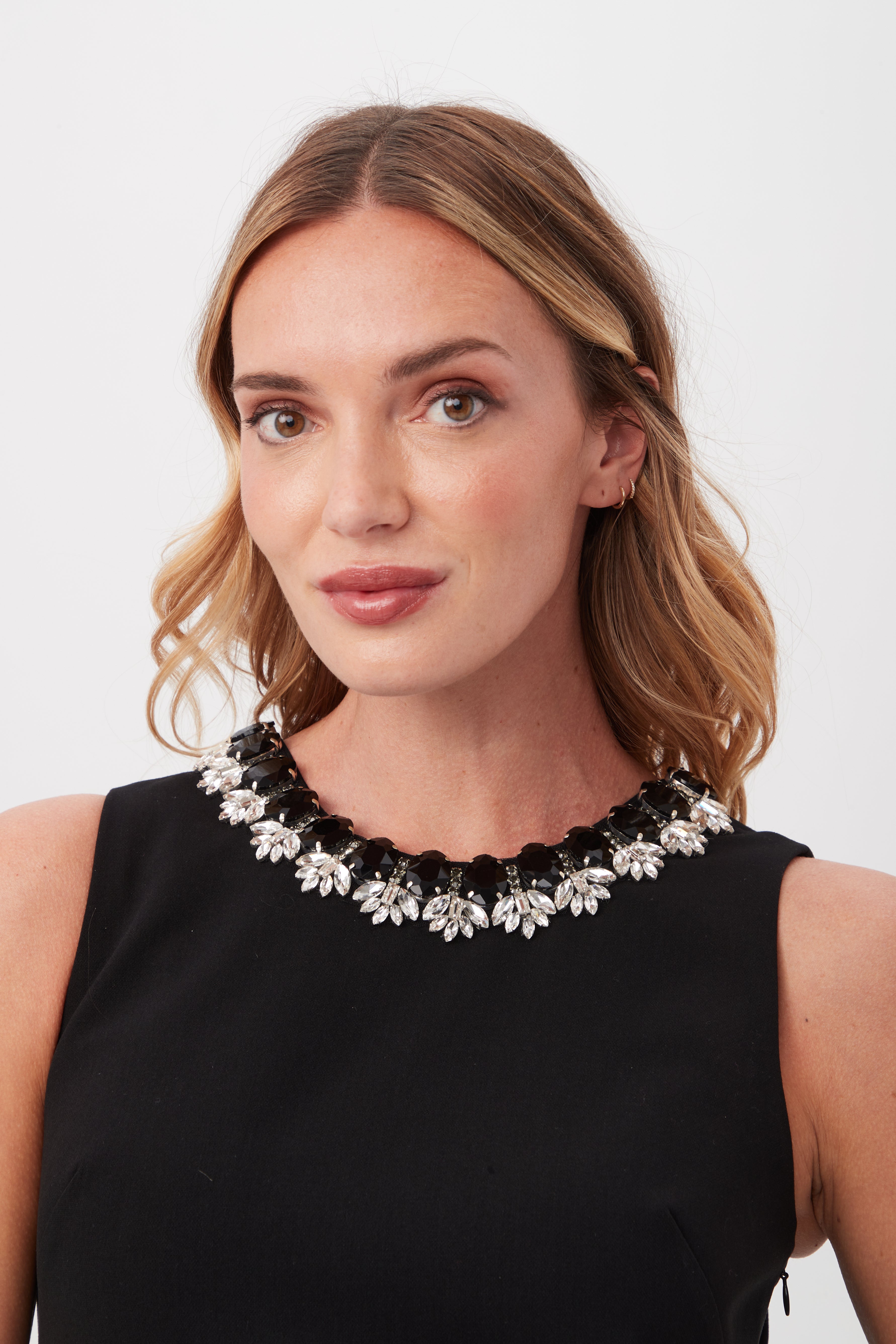 A woman with shoulder-length blonde hair models the Beauty 2 Black Short Sleeveless A-Line Dress and a statement necklace with silver and black floral accents, posing against a plain light background.