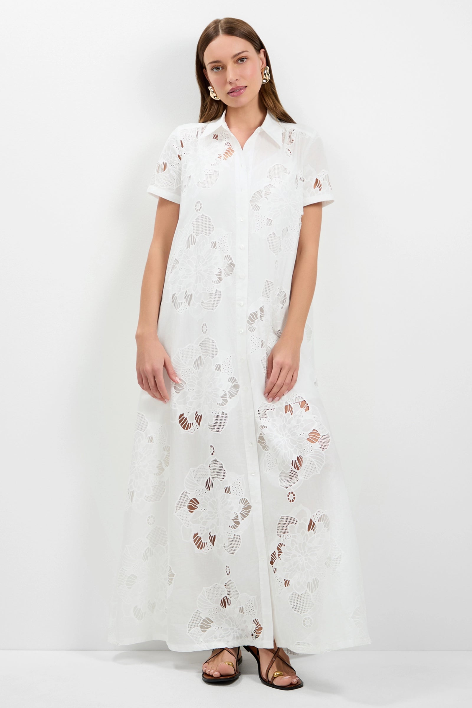 A woman with long brown hair wears the Kon Tiki Maxi Dress in Whitewash—a long, white short-sleeve dress with sheer floral patterns and front buttons. She pairs it with gold earrings and brown sandals, standing against a white background.