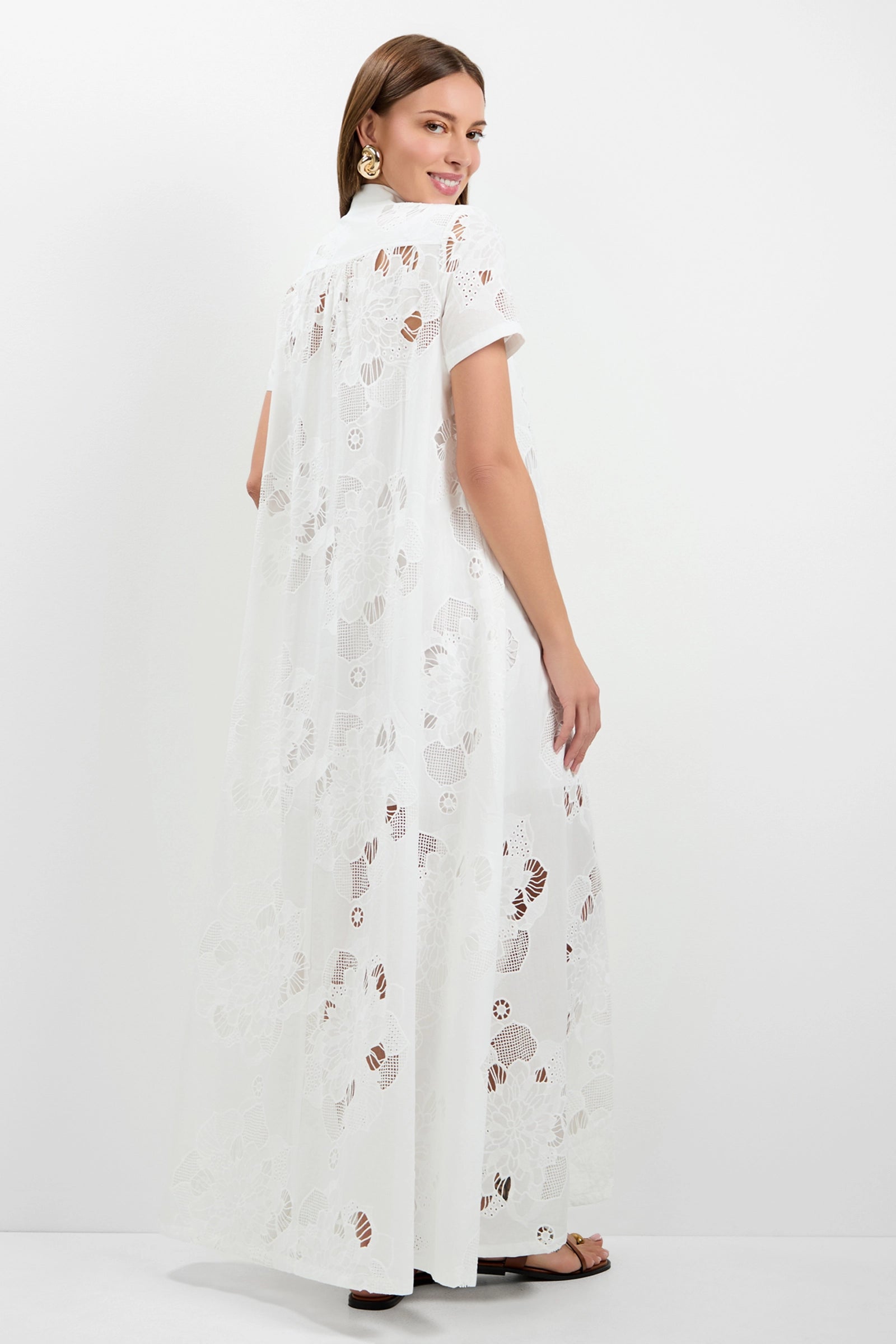 A woman with straight brown hair, gold earrings, and black sandals stands against a white background, looking over her shoulder and smiling while wearing the Kon Tiki Maxi Dress—a long, white, sheer floral lace dress with short sleeves.