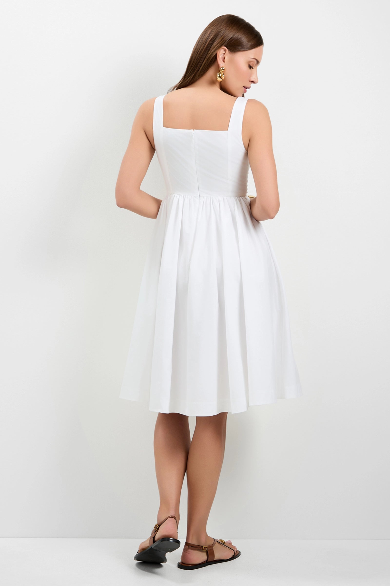 A woman with straight brown hair and gold earrings wears the Mirabello Sweetheart Neck Midi Dress, a sleeveless white fitted-bodice, flared skirt midi, paired with brown flat sandals. She stands facing away against a plain white background.