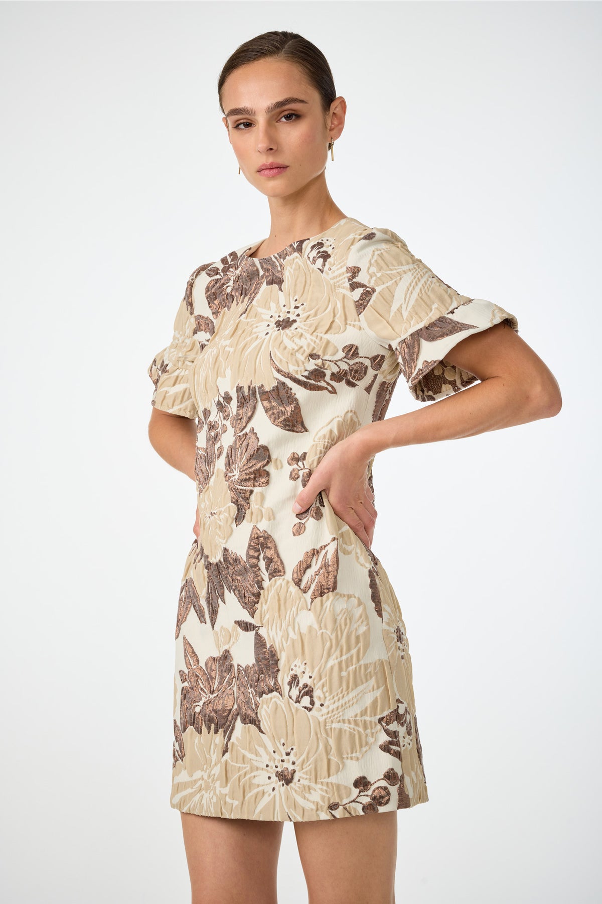 A woman poses in the Darling Flounce Sleeve Dress, featuring cream fabric with brown and beige floral patterns, short sleeves, and standing against a plain white background with her hands on her hips.