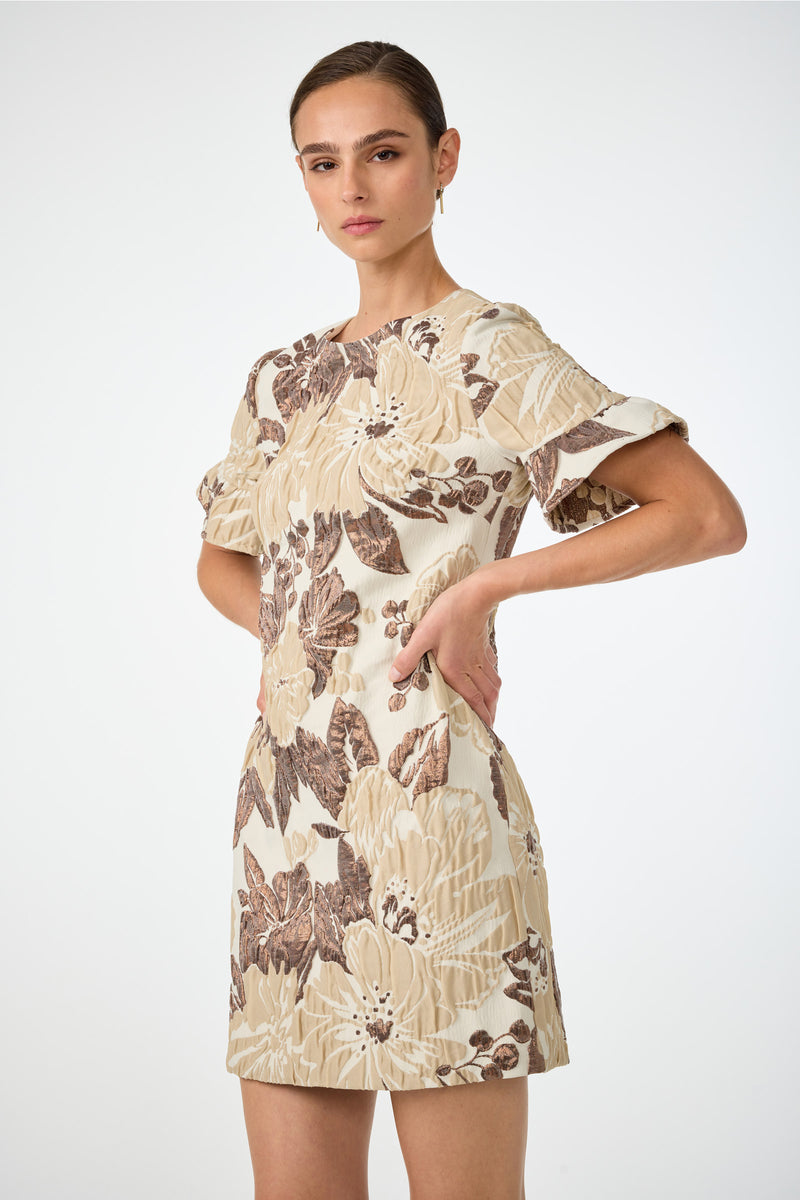 A woman poses in the Darling Flounce Sleeve Dress, featuring cream fabric with brown and beige floral patterns, short sleeves, and standing against a plain white background with her hands on her hips.