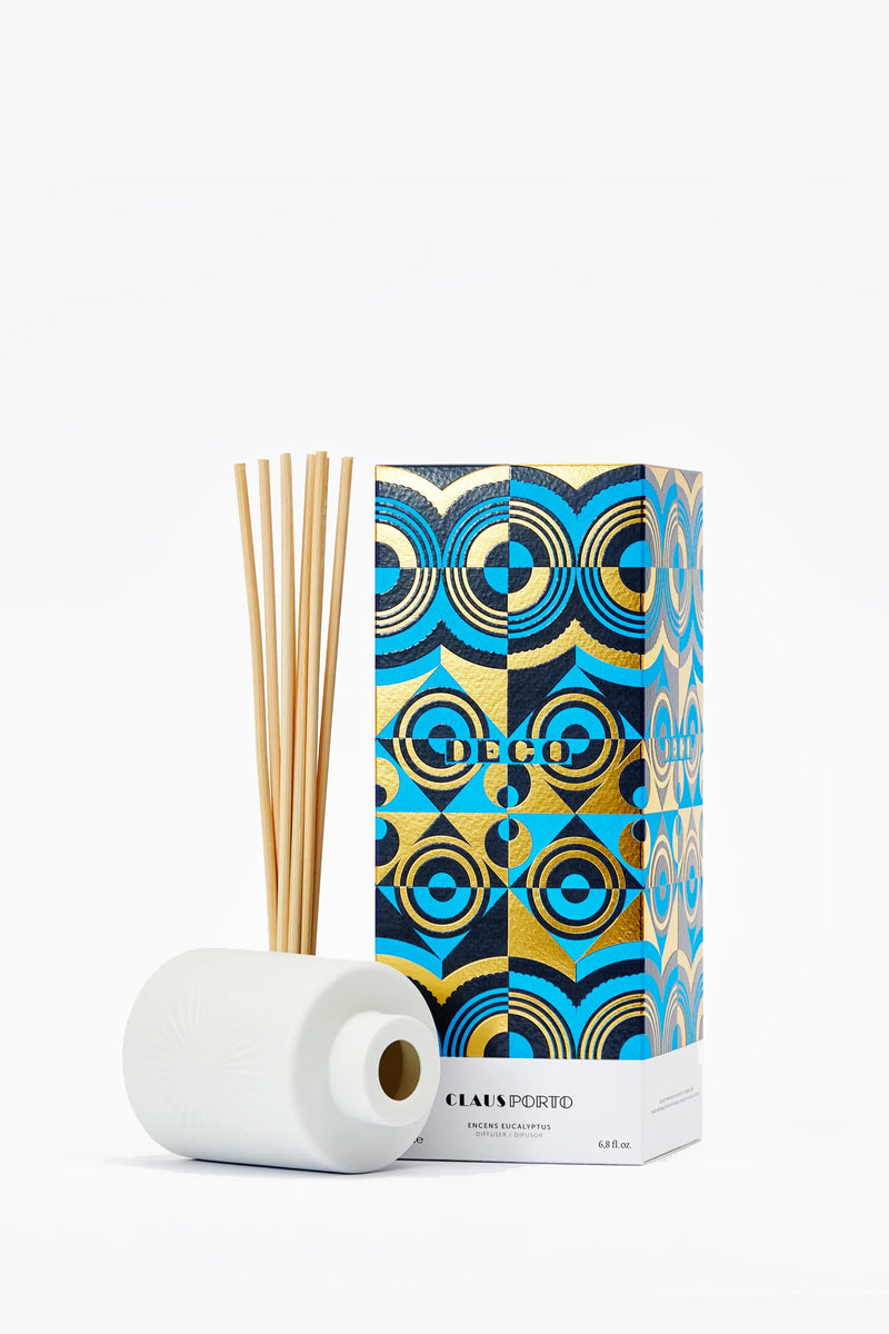 The Claus Porto Deco Diffuser, featuring a white bottle with wooden reeds, stands beside its colorful box adorned with blue, gold, and black geometric patterns. The box displays the Claus Porto name against a white background.