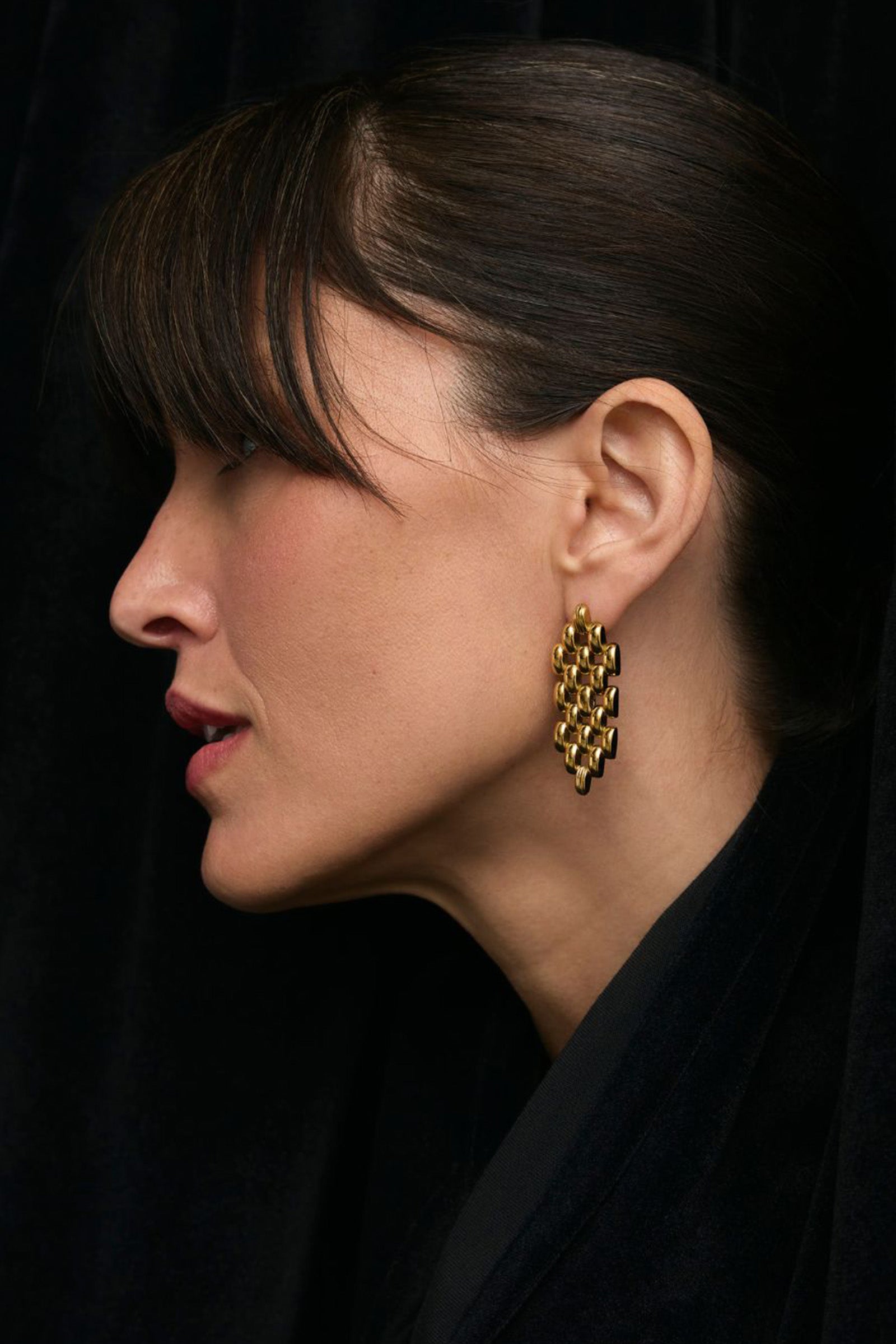 A woman with dark hair and bangs, dressed in black, is shown in profile against a dark background. She wears gold geometric Phoebe Earrings.