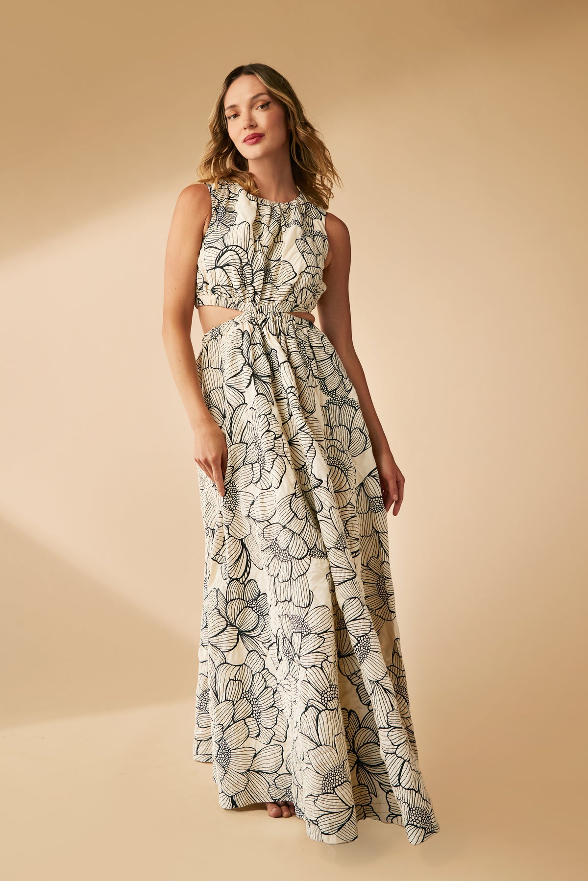 A woman models the Artimo Maxi Dress—a sleeveless, floor-length cream dress with bold black floral prints and waist side cutouts—while posing against a beige backdrop.