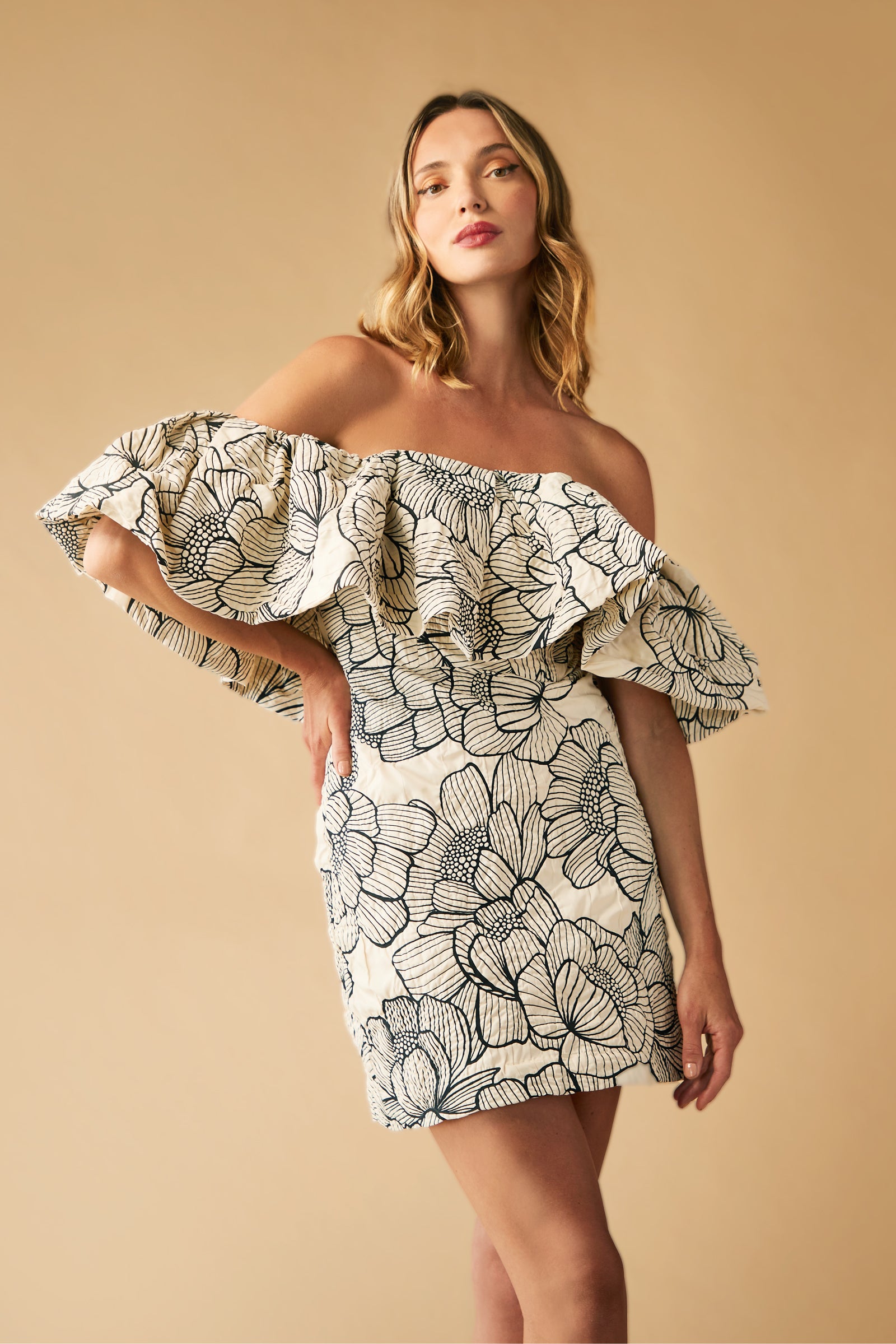 A woman poses in the Nia Dress—an off-the-shoulder, ruffled mini dress with a bold black floral print on cream—standing against a beige backdrop.