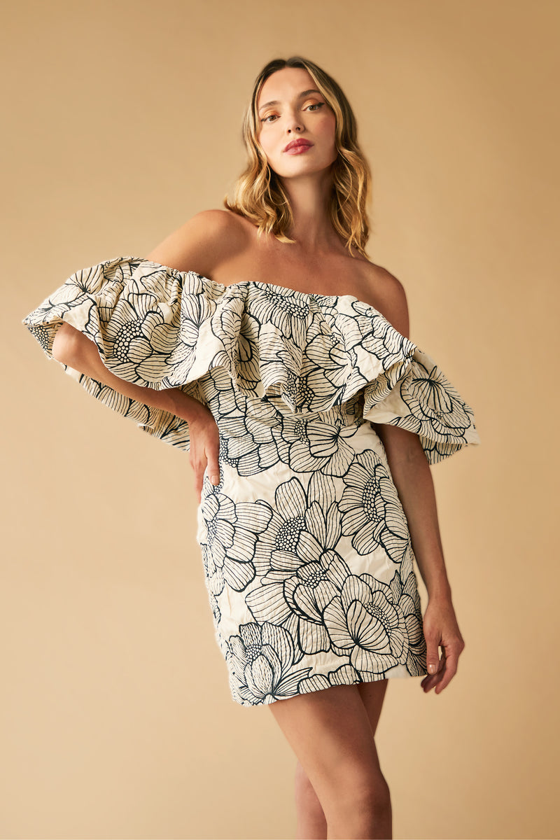 A woman poses in the Nia Dress—an off-the-shoulder, ruffled mini dress with a bold black floral print on cream—standing against a beige backdrop.
