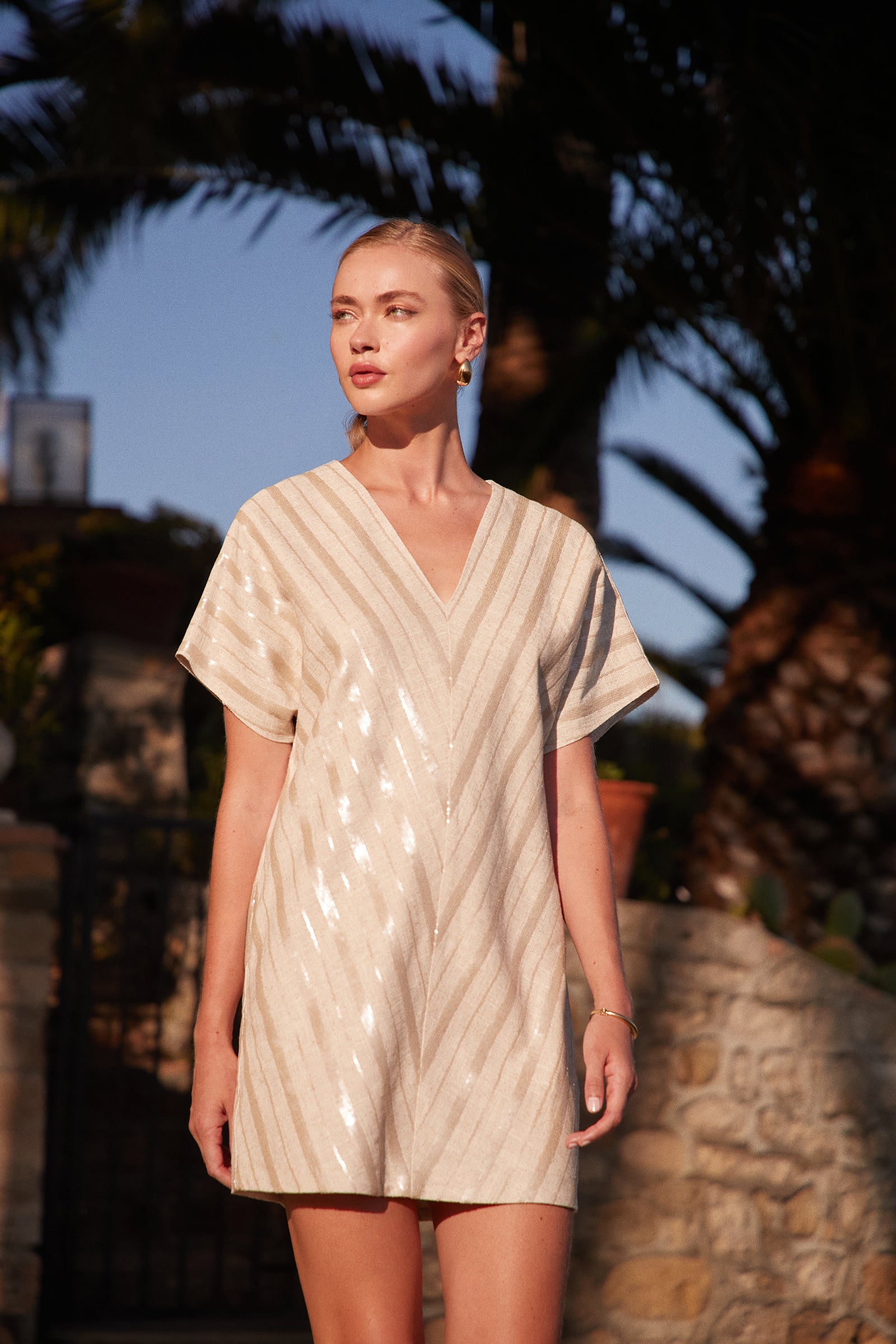 A woman stands outdoors in sunlight, wearing the Elisabetta Short-Sleeve Dress, a cream-colored, V-neck style with diagonal shiny stripes. Palm trees and stone structures appear in the background.