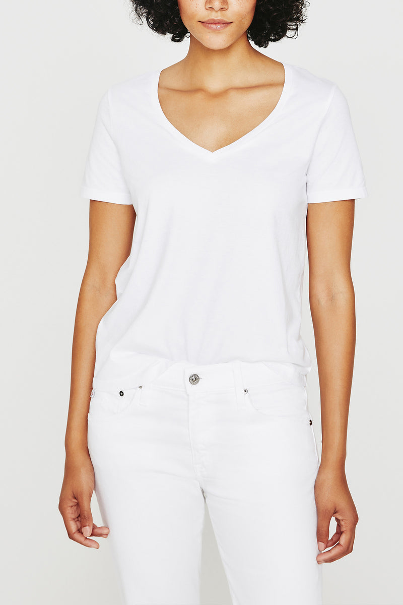 A woman poses against a plain background wearing the AG Jager Vee Tee in white, paired with white jeans. Her head is slightly tilted, and only her upper body is shown.