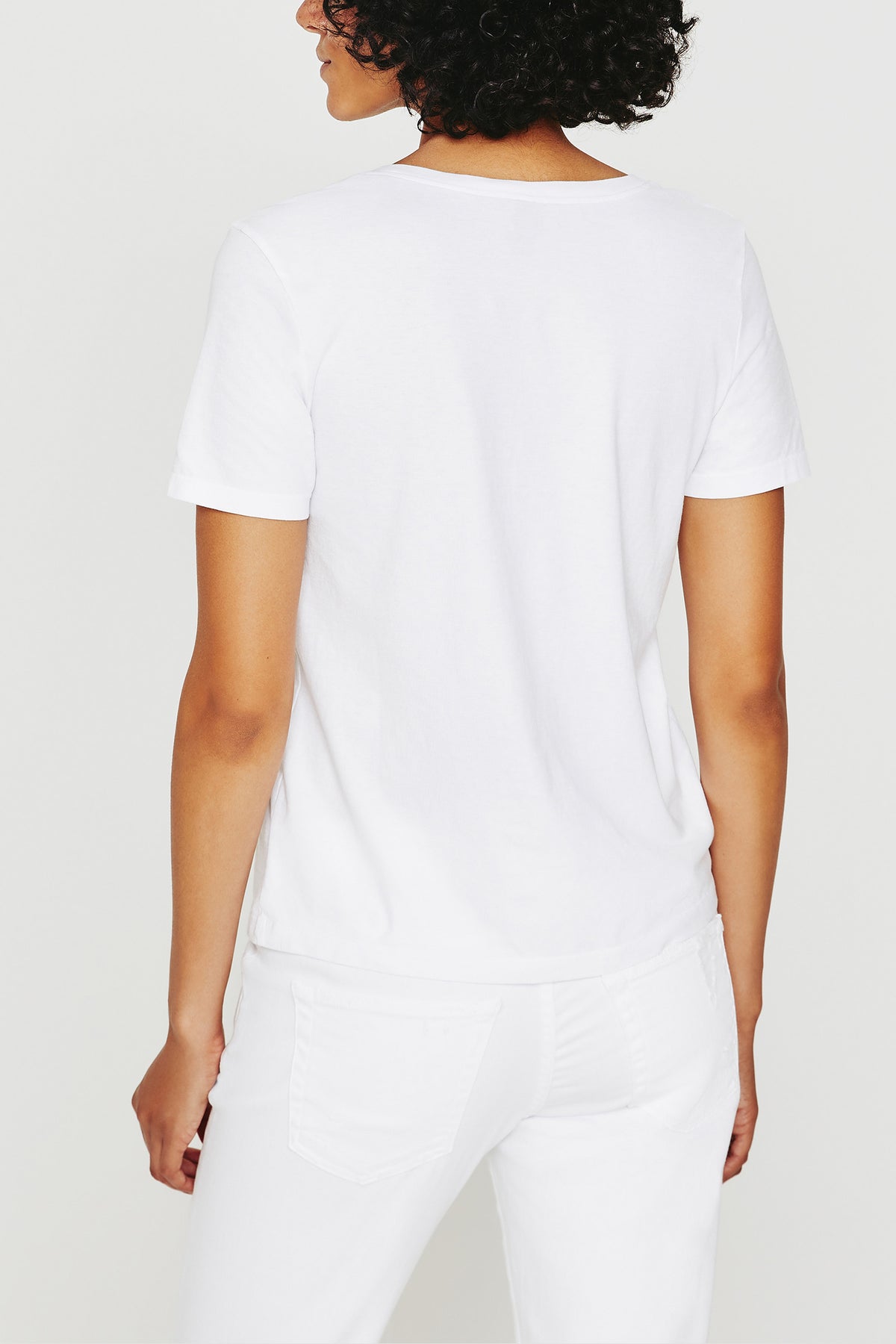 A person with short curly hair is shown from the back, wearing the AG Jager Vee Tee in white and white pants, standing against a plain white background.