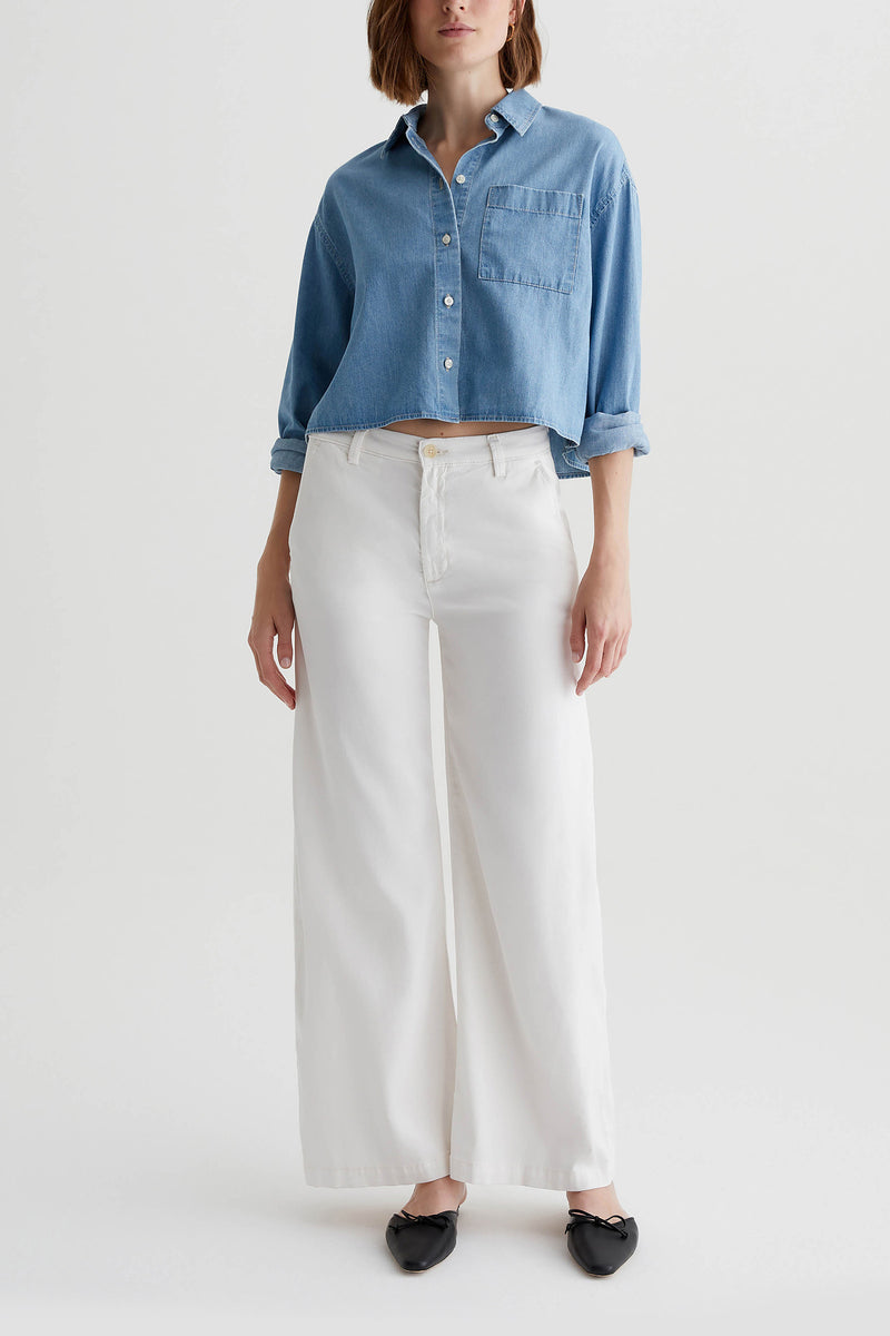 A woman stands against a white background wearing a light blue cropped shirt and the AG Caden Wide Leg Pant in Powder, along with black pointed-toe flats. The pants feature a wide-leg silhouette for a modern look.