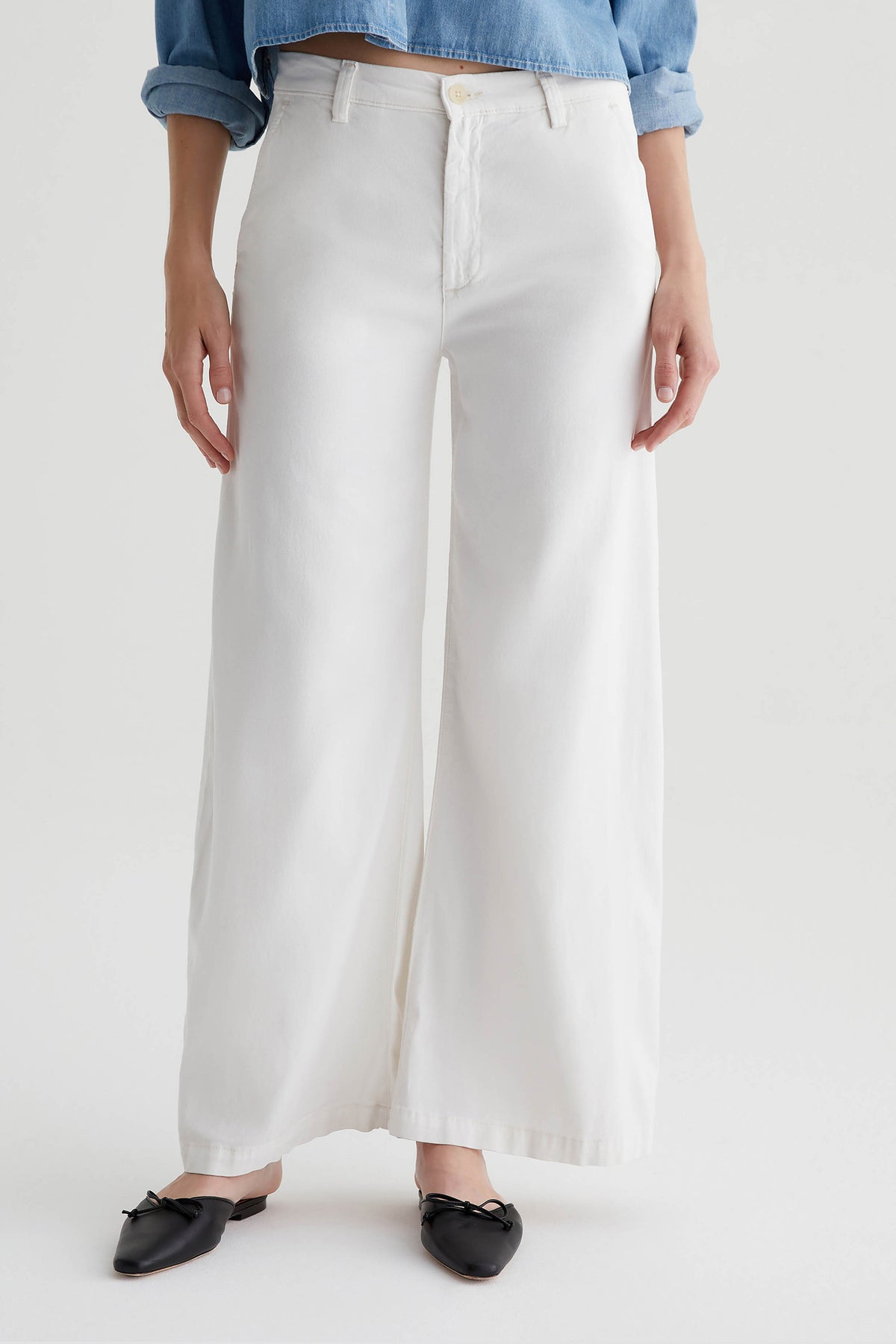 A person wearing the AG Caden Wide Leg Pant in Powder, paired with a light blue rolled-sleeve shirt and black pointed-toe flats, stands against a plain white background.