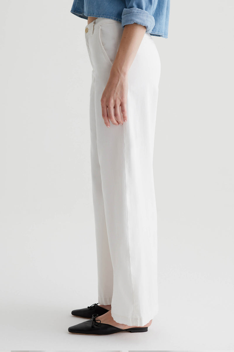 A person stands in profile wearing the AG Caden Wide Leg Pant in Powder, a light blue rolled-sleeve shirt, and black flat shoes with a small bow. The background is plain and light.