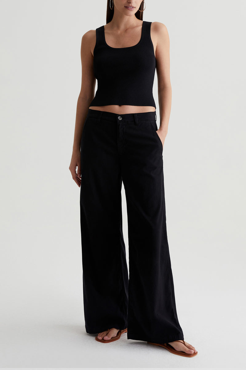A person stands against a light background wearing the AG Caden Wide Leg Pant in black, paired with a black sleeveless crop top and brown flip-flops. Their hands are in their pockets, and their face is not visible.