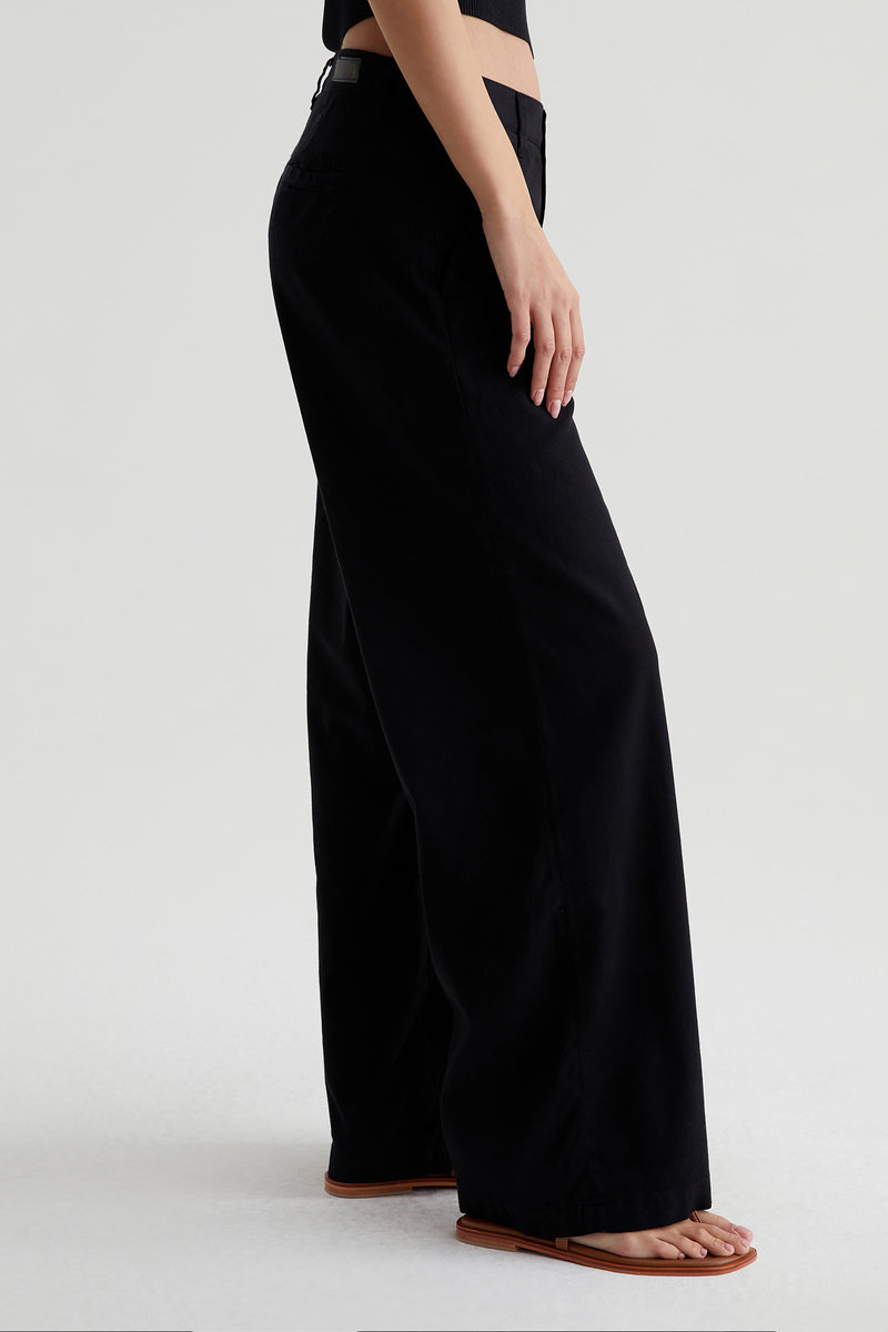 A person wearing the AG Caden Wide Leg Pant in Black and flat brown sandals, shown from the waist down against a plain, light background.