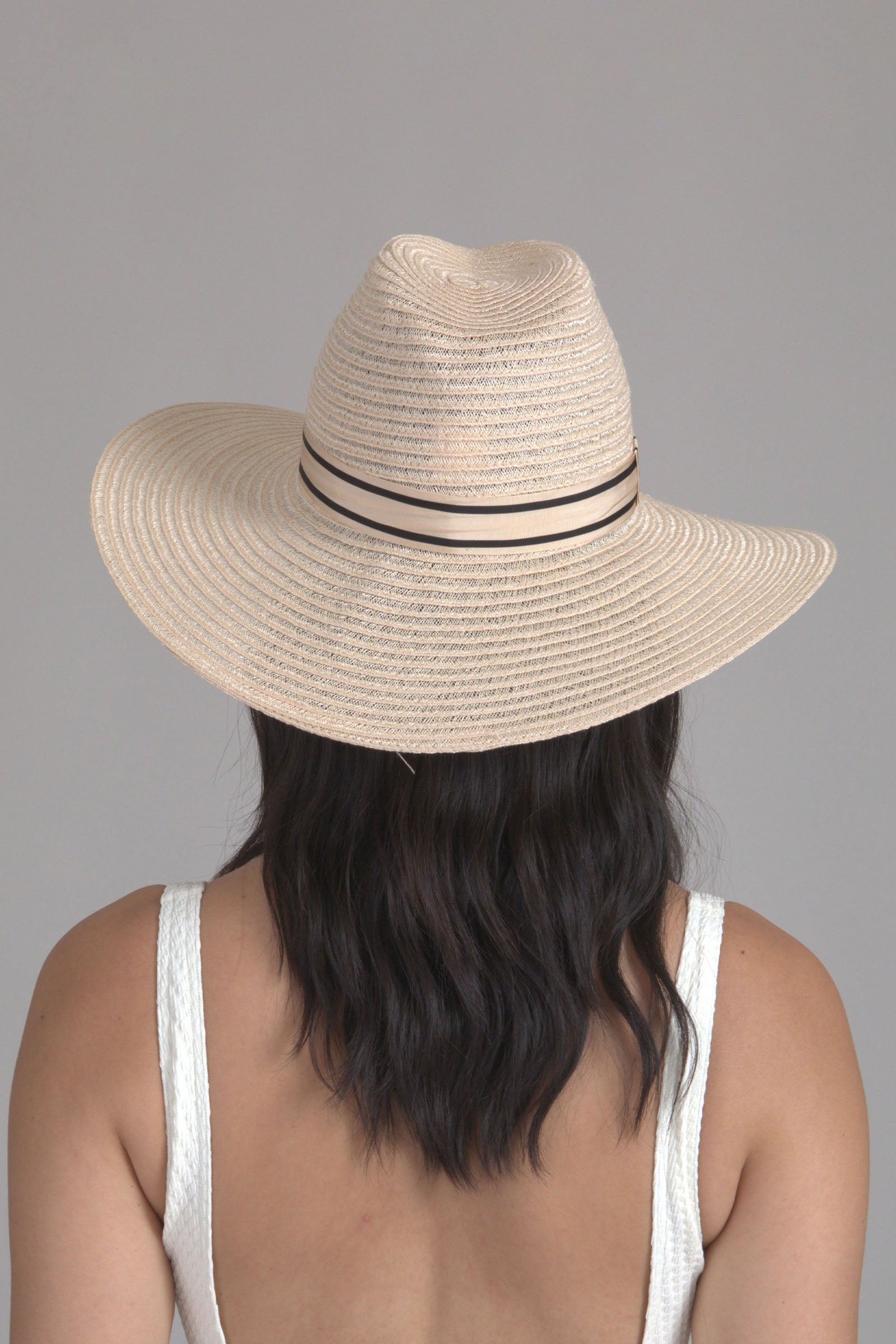 A person with wavy dark hair is seen from behind, wearing the Eugenia Kim Emmanuel Sun Hat. The wide-brimmed straw hat with a thin ribbon frames their white textured sleeveless top against a plain background.