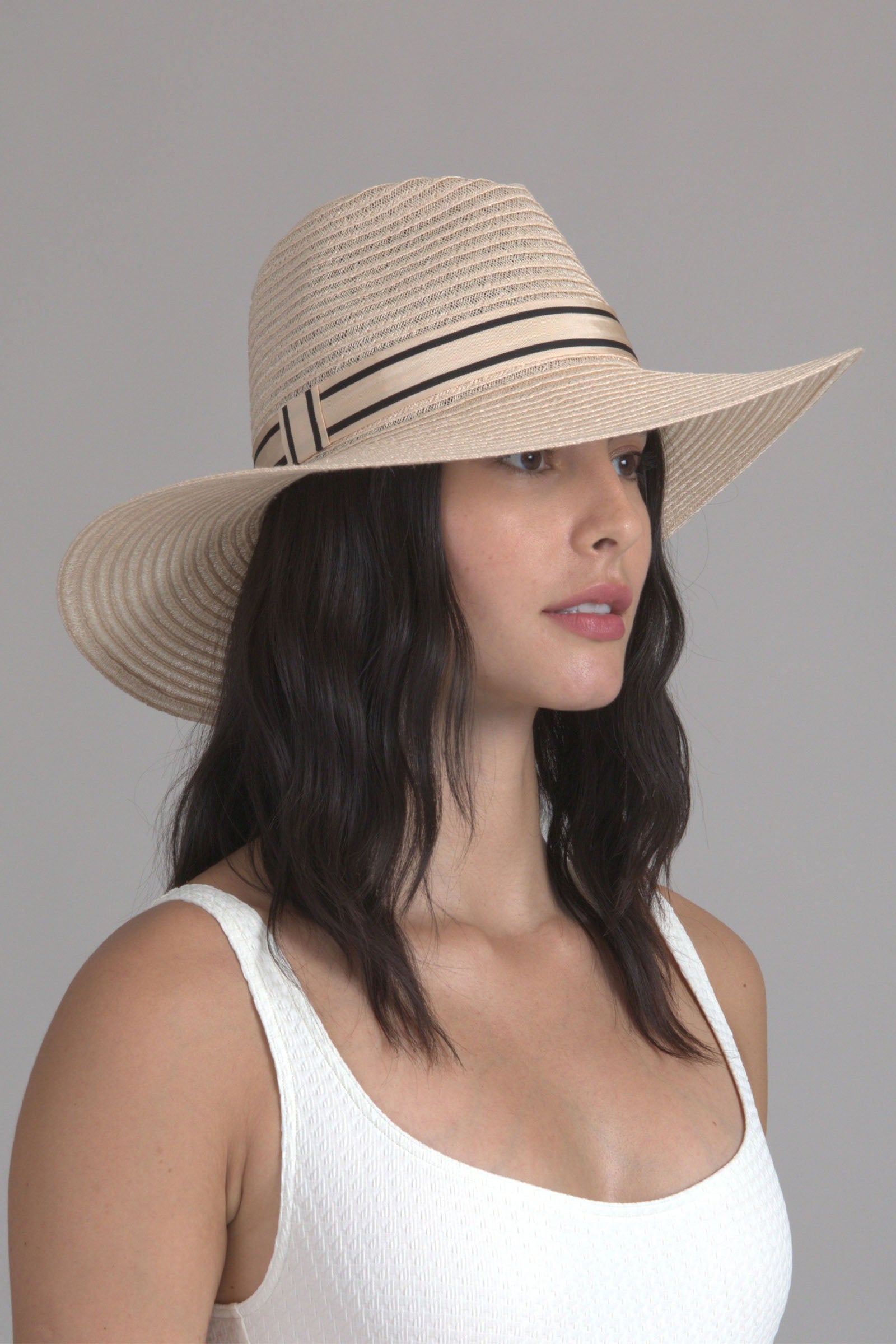 A woman with long dark hair wears the Eugenia Kim Emmanuel Sun Hat, featuring a wide brim and black and cream ribbon, paired with a white textured tank top, posing against a light gray background.