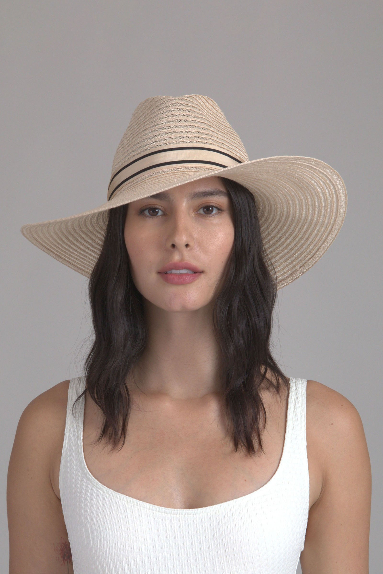 A woman with wavy dark hair wears the Eugenia Kim Emmanuel Sun Hat, featuring a wide woven straw brim and black band, paired with a white textured sleeveless top against a plain light gray background.