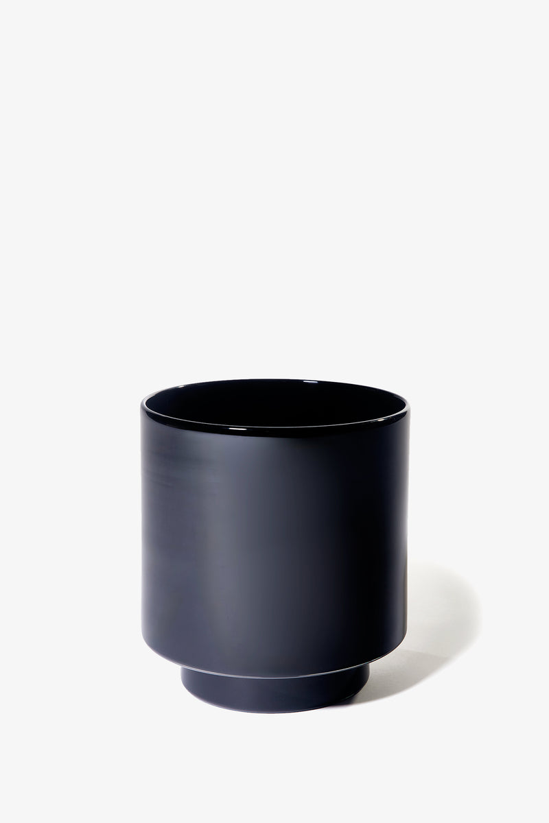 The Fiorelli Low Container is a minimalist, cylindrical black ceramic planter with a small elevated base, set against a plain white background.