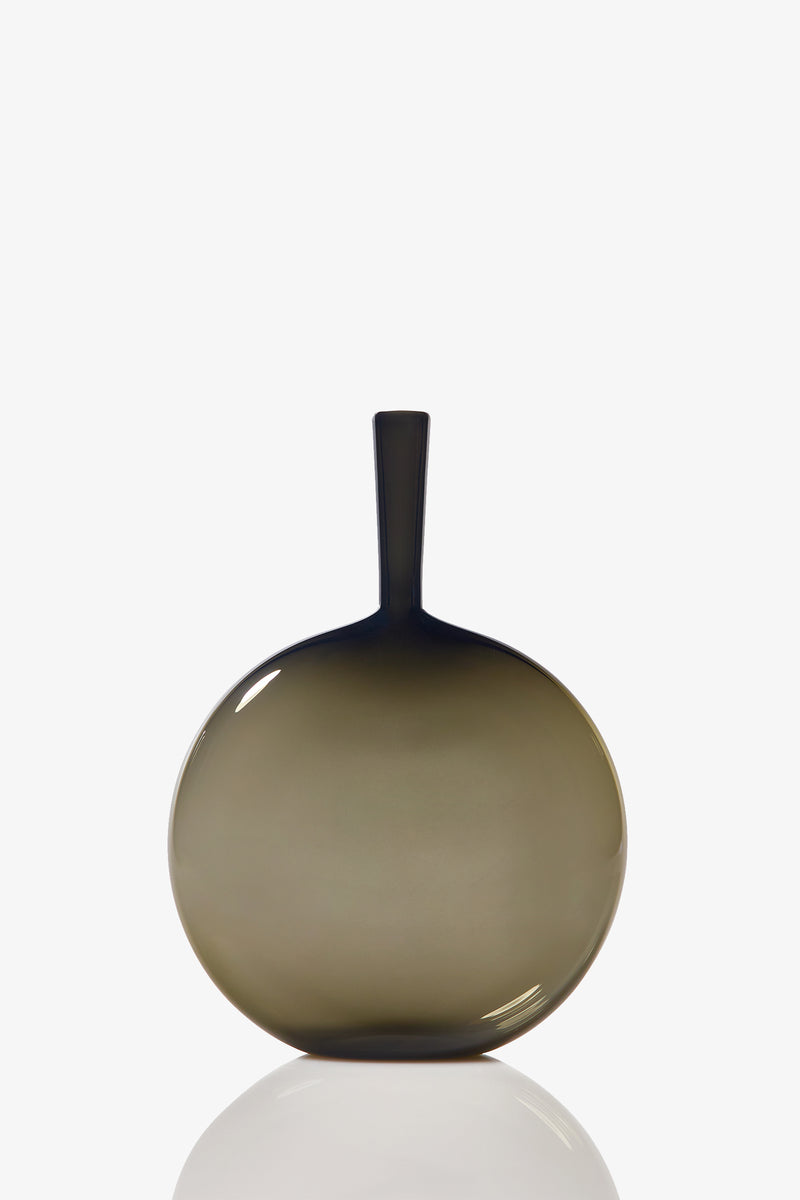 The Flask Angelic Bottle is a round, smoky gray glass vase with a flat body and long, narrow neck. It features a smooth, glossy finish and casts a subtle reflection against a plain white background.
