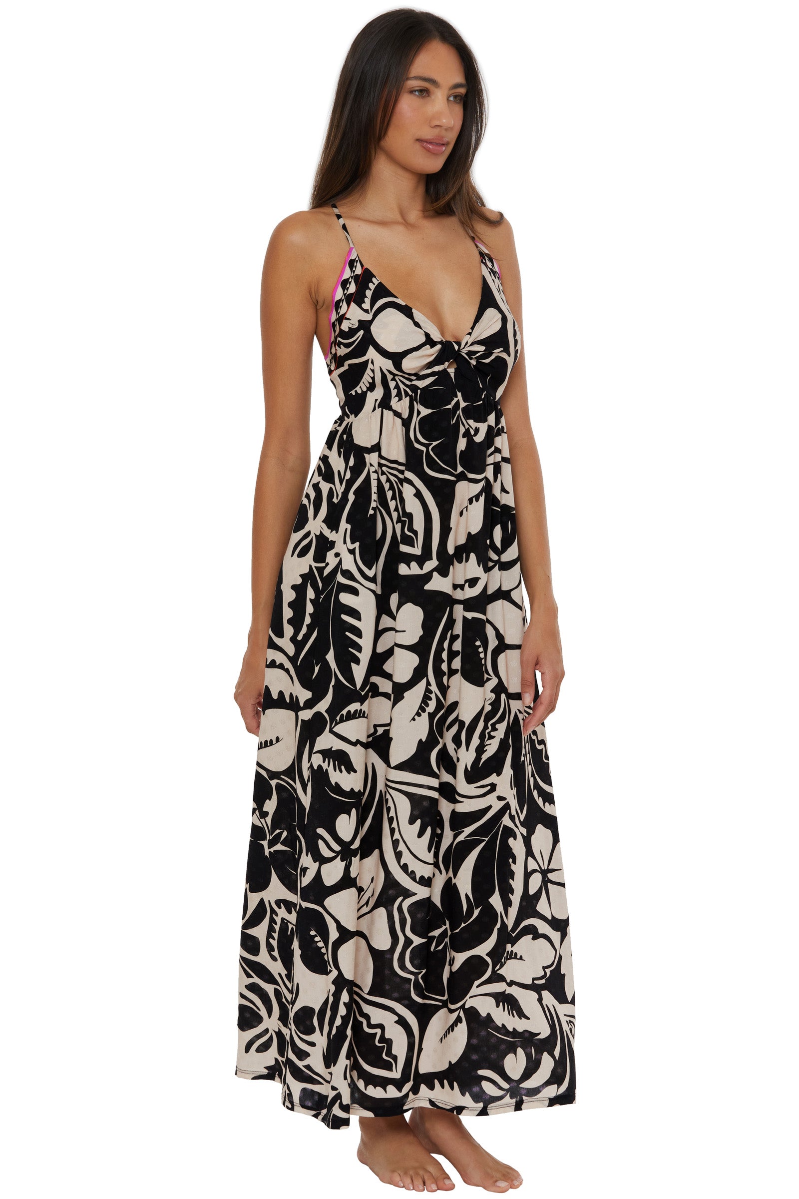 A woman with long brown hair and a relaxed expression is barefoot, wearing the Twist Back Maxi Dress Cover-Up - Florence, featuring a bold black and beige tropical leaf print, sleeveless design, deep V-neck, and maxi length.