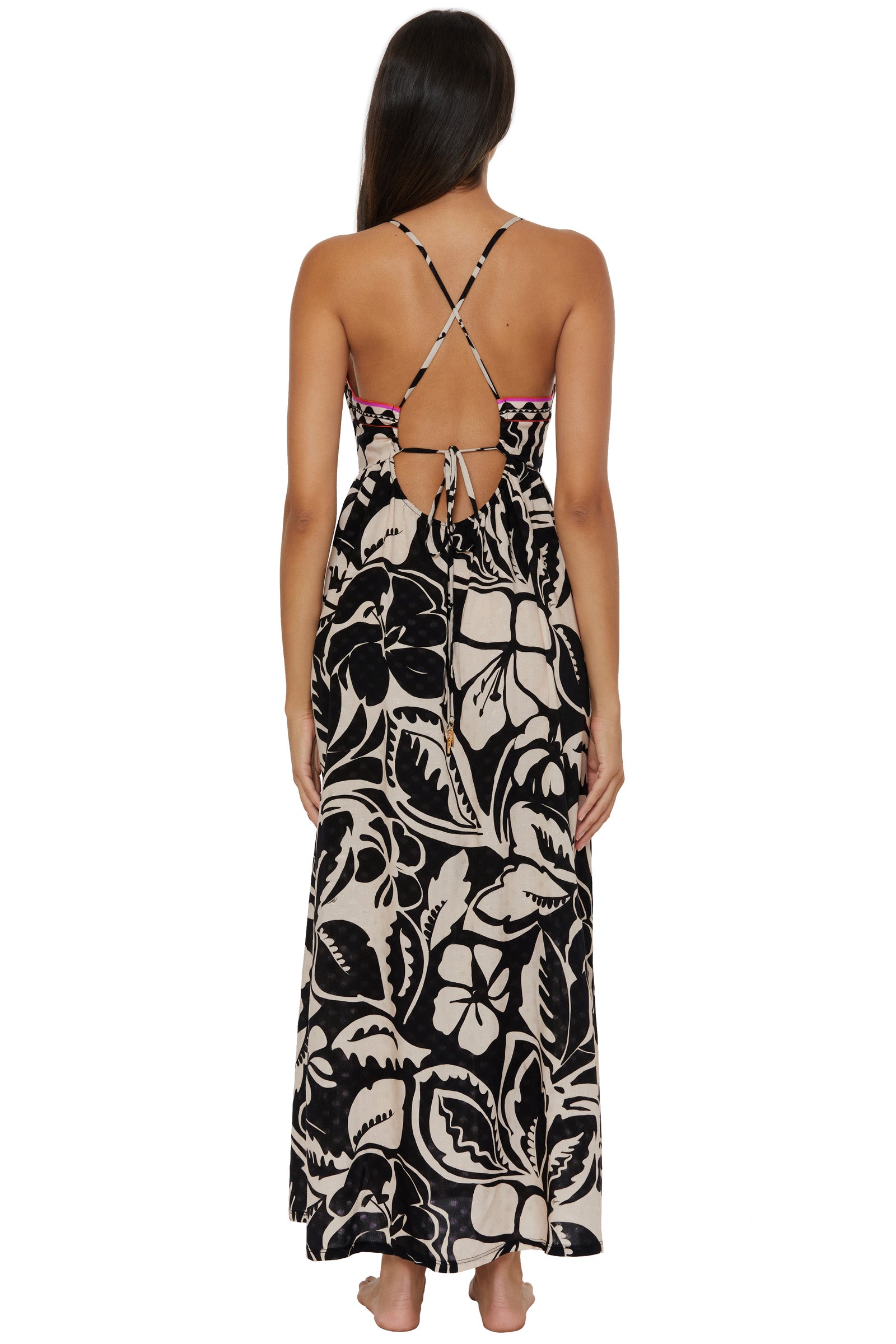 A woman stands barefoot, facing away, wearing the Twist Back Maxi Dress Cover-Up - Florence, a long black and white floral halter dress with crisscross straps and an open back.