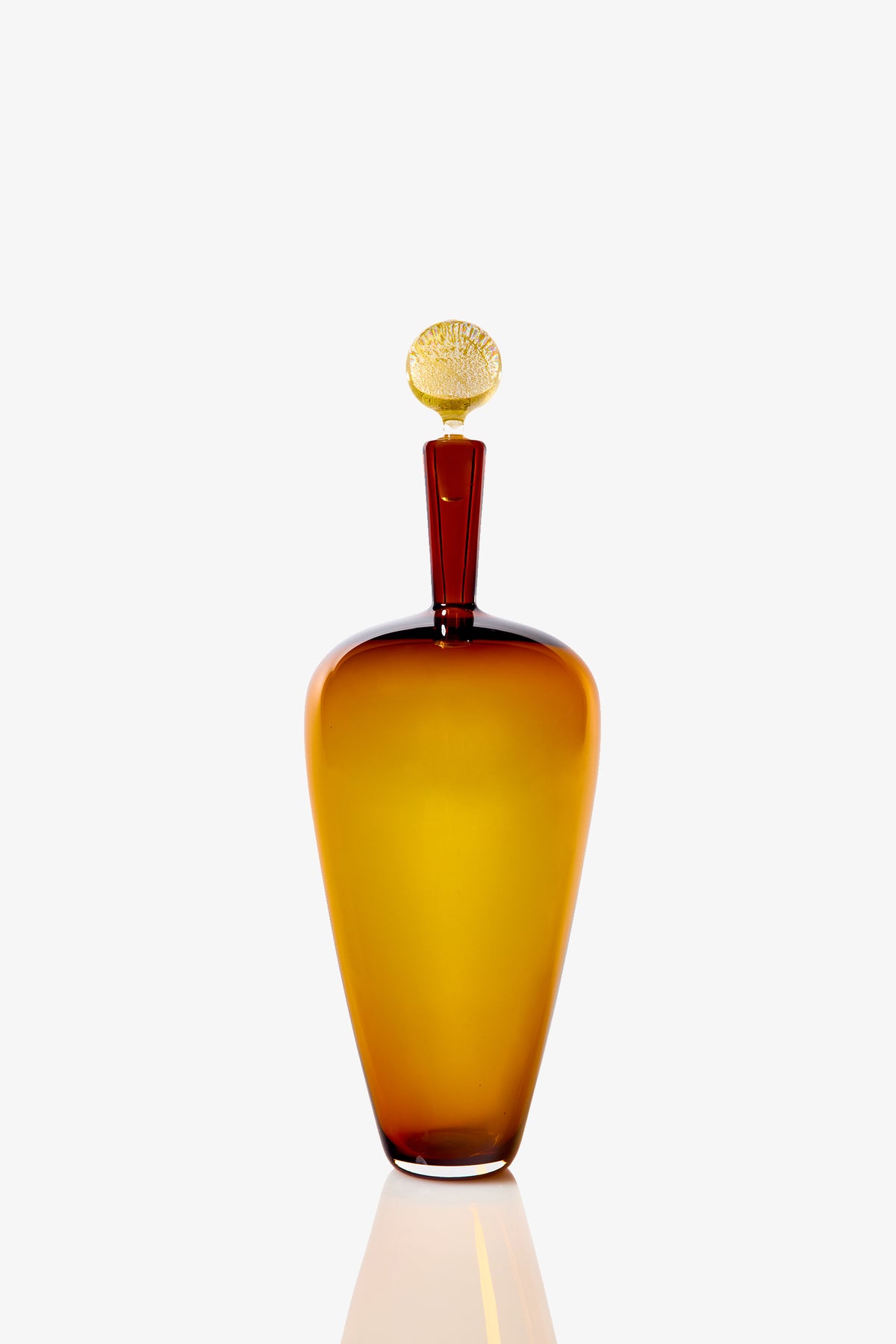 The High Shoulder Carmella Barware features a tall, elegant amber glass bottle with a rounded body and matching stopper, standing upright on a white reflective surface against a plain white background.
