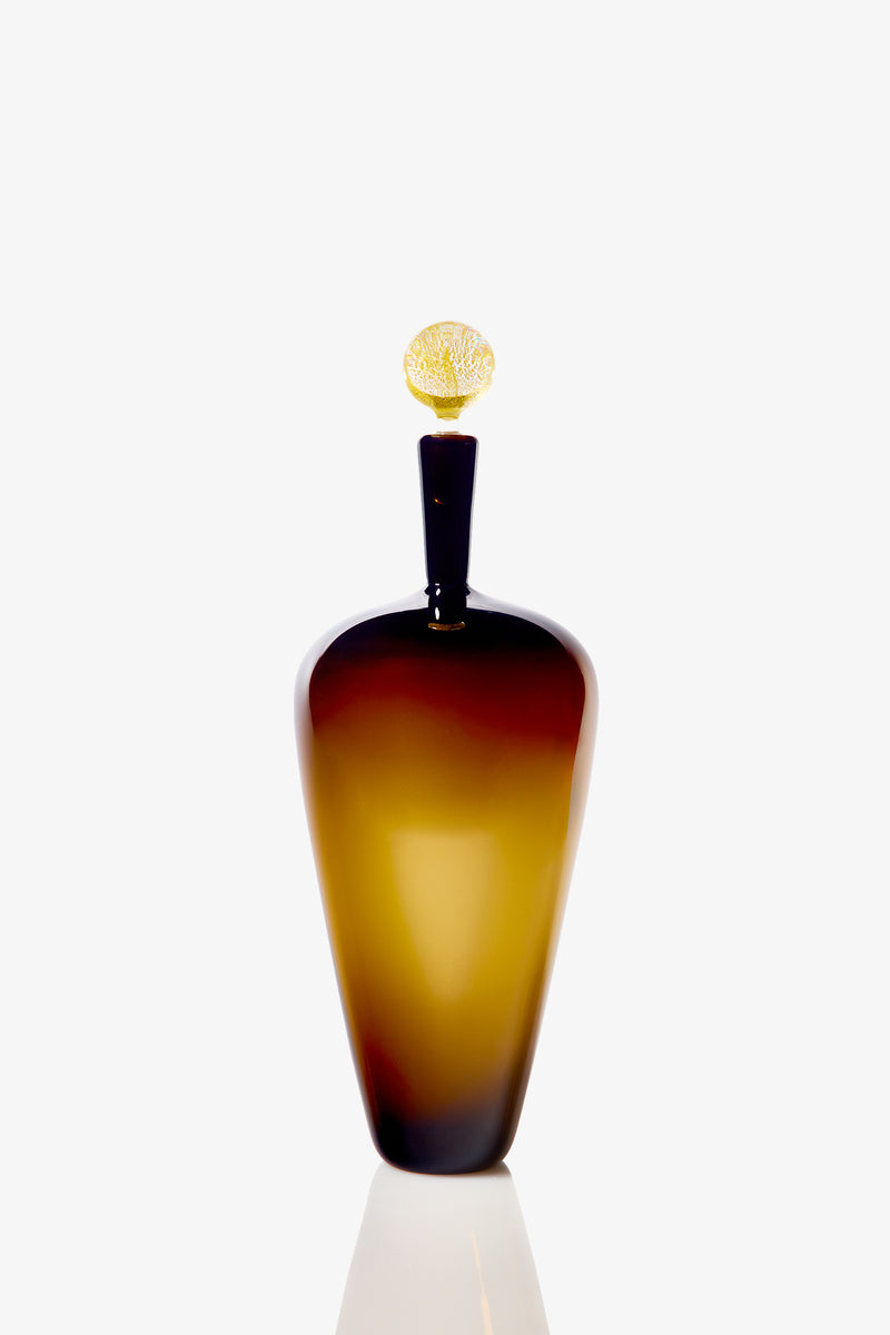 The High Shoulder Carmella Barware features a tall, elegant amber glass design with a rounded body, narrowed neck, and a textured golden stopper, beautifully presented against a plain white background.