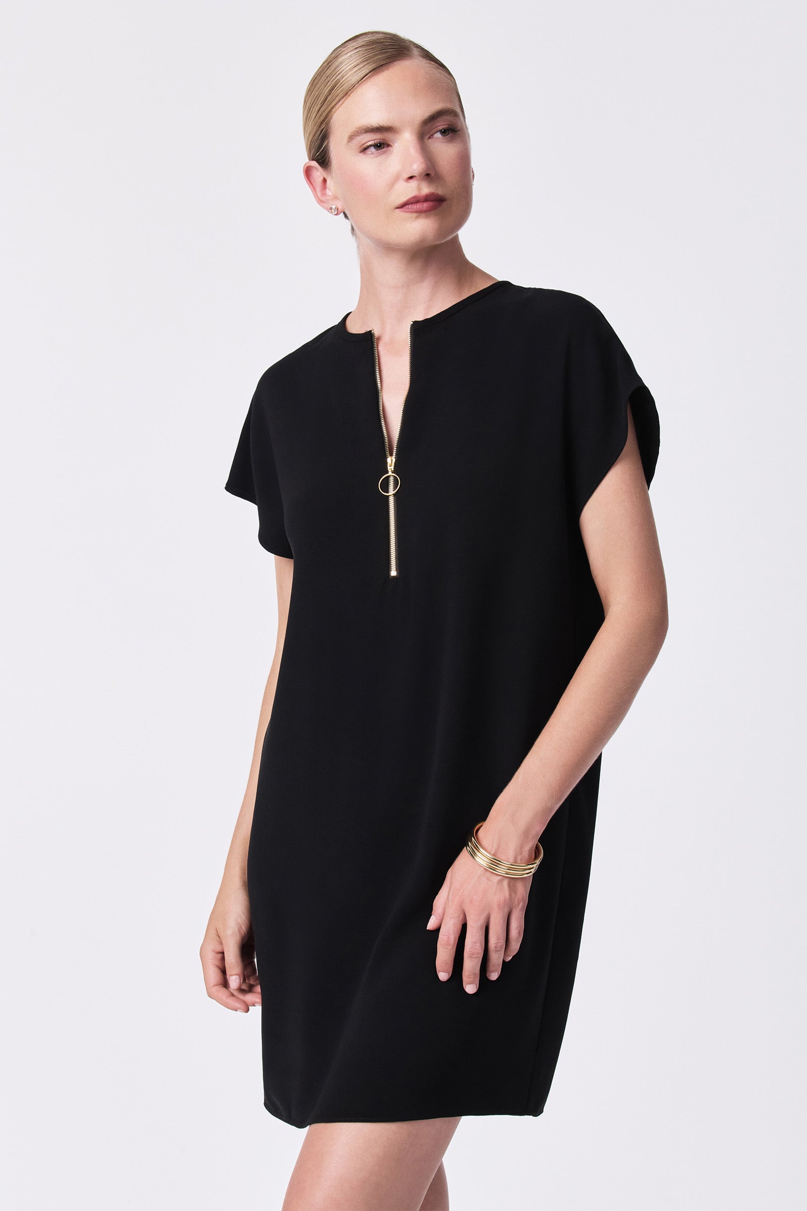 A woman wears the Hasil Short-Sleeve Shift Dress, featuring a front zipper detail, standing against a plain white background with her hair pulled back and gold bracelets as accessories.