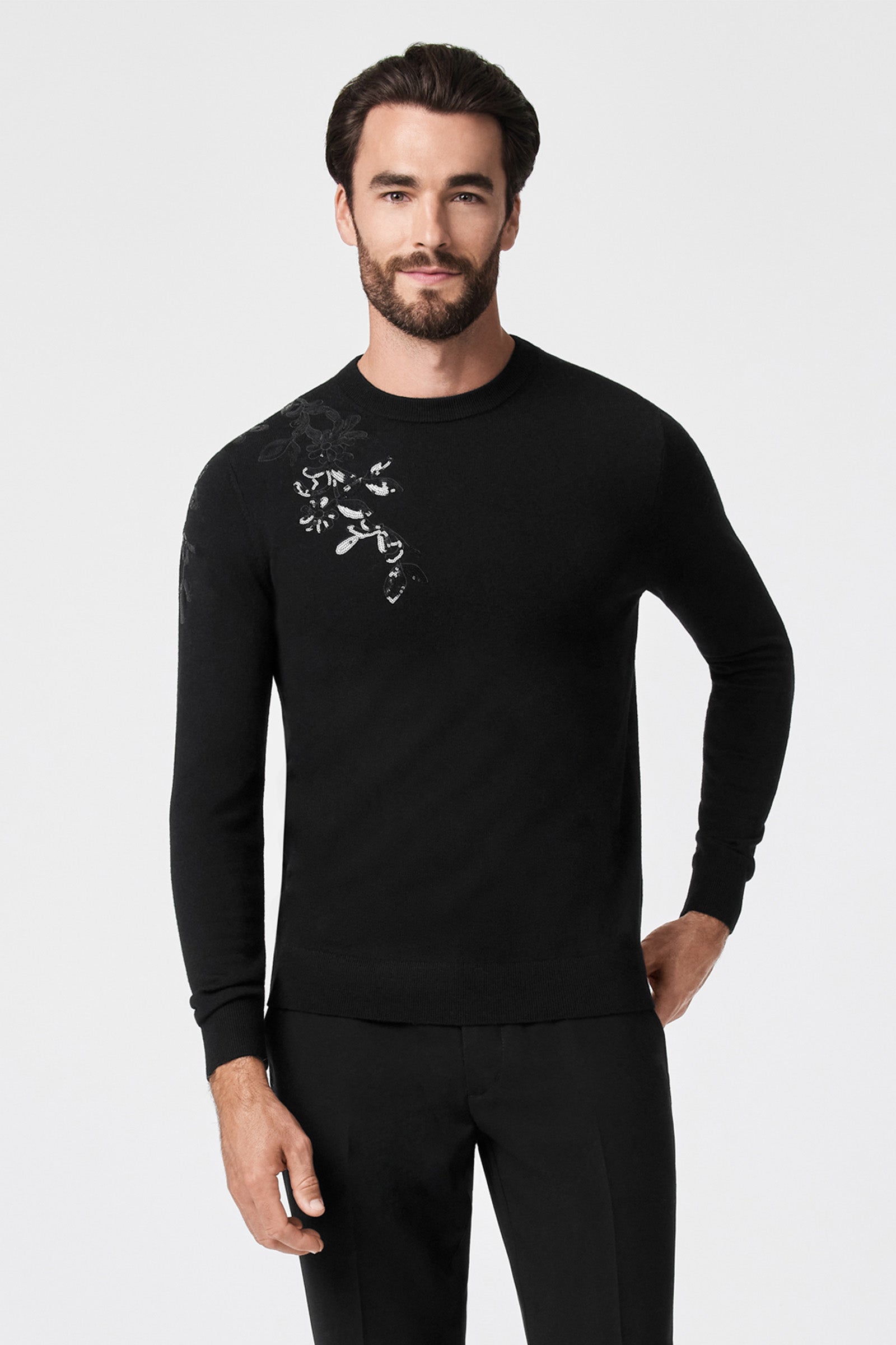 A man with dark hair and a beard wears the Jaxon Sequin Crewneck Sweater, featuring a subtle floral design on the left shoulder, paired with black pants. He stands against a plain white background, looking at the camera with a slight smile.