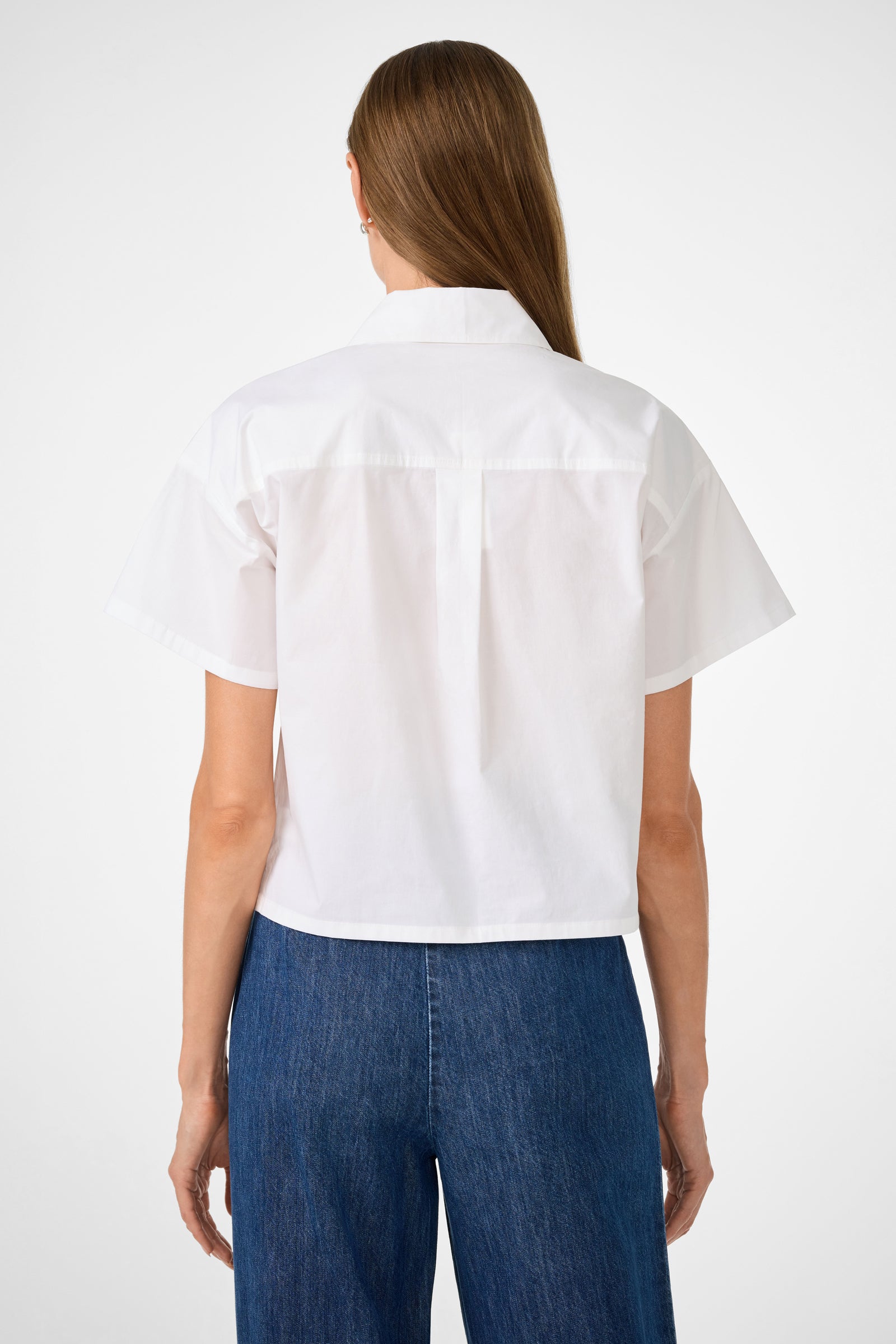 A person with straight, light brown hair is shown from the back, wearing the Komore Cropped Button-Front Top in white and blue jeans against a plain, light background.
