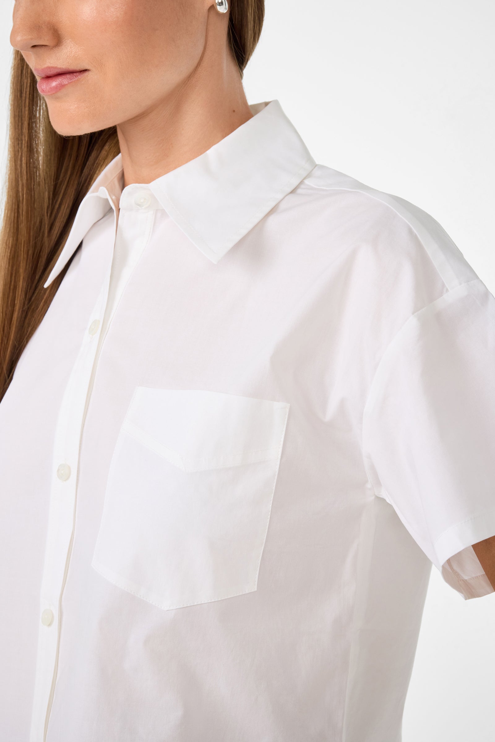 A woman wears the Komore Cropped Button-Front Top, a white short-sleeve shirt with a front pocket, shown from neck to mid-arm against a plain white background.