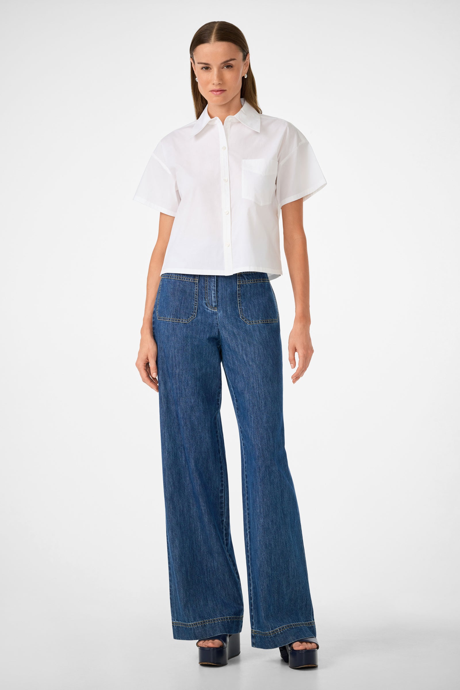 A woman stands against a plain background, wearing the Komore Cropped Button-Front Top, short-sleeve and white, paired with high-waisted wide-leg blue jeans featuring large front pockets and black platform sandals.