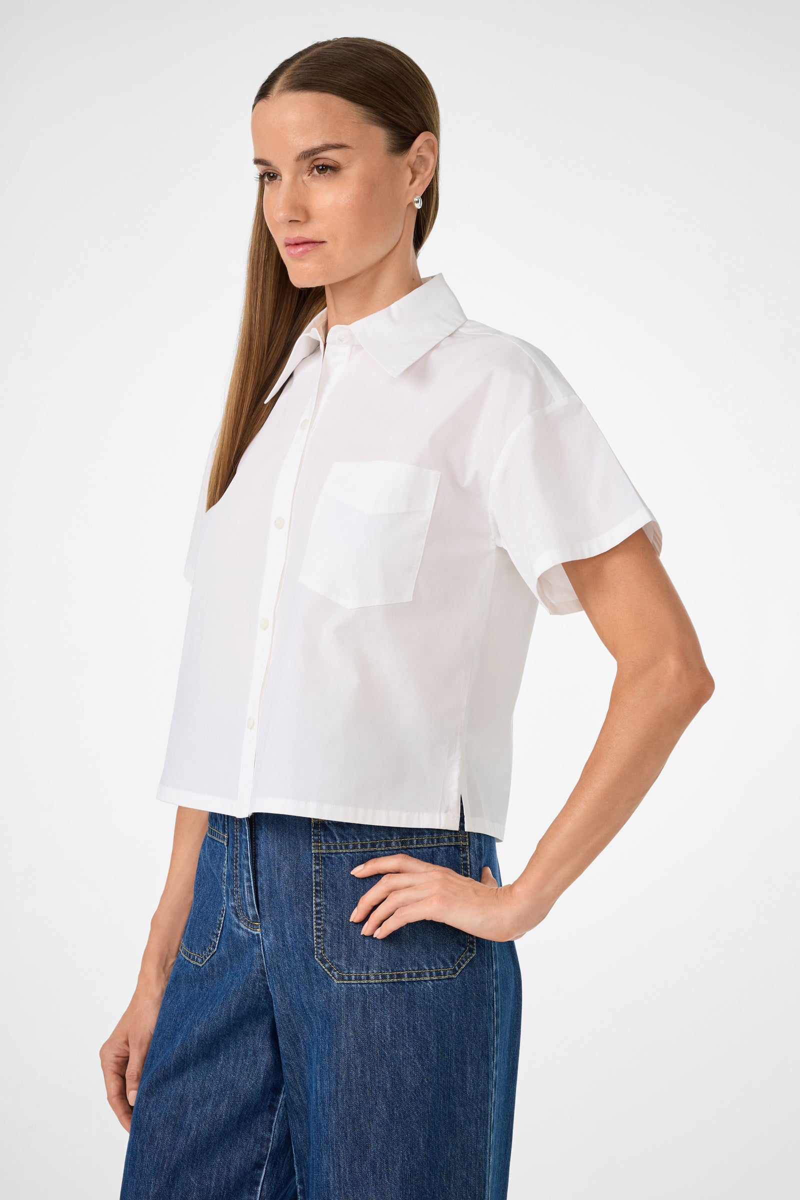 A woman with straight brown hair models the Komore Cropped Button-Front Top, a white short-sleeved shirt with a chest pocket, paired with high-waisted blue jeans. She poses with one hand on her hip against a plain background.