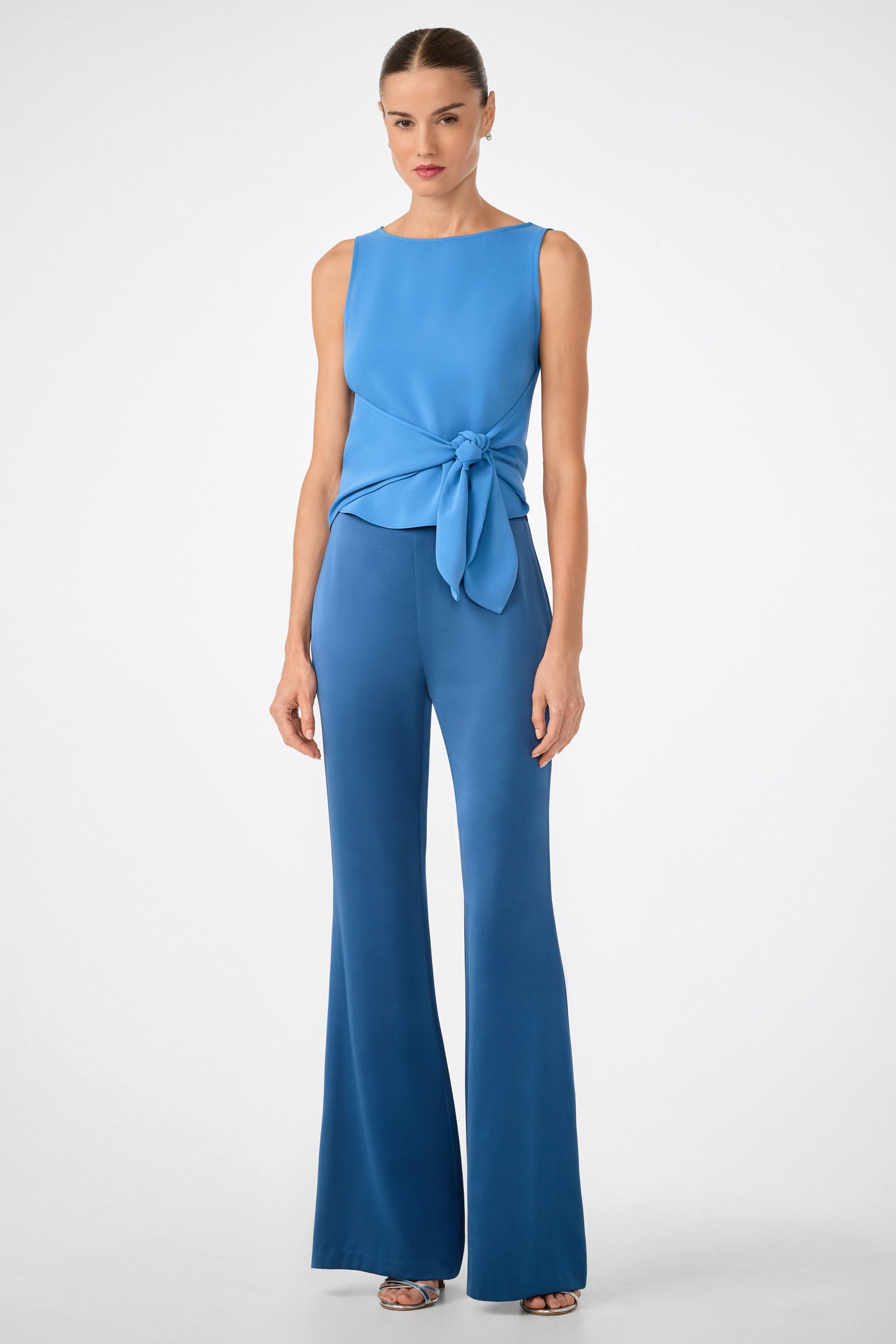 A woman in the Kuri Tie-Front Top, a sleeveless light blue waist-tie top, paired with matching flared blue pants stands against a plain white background.