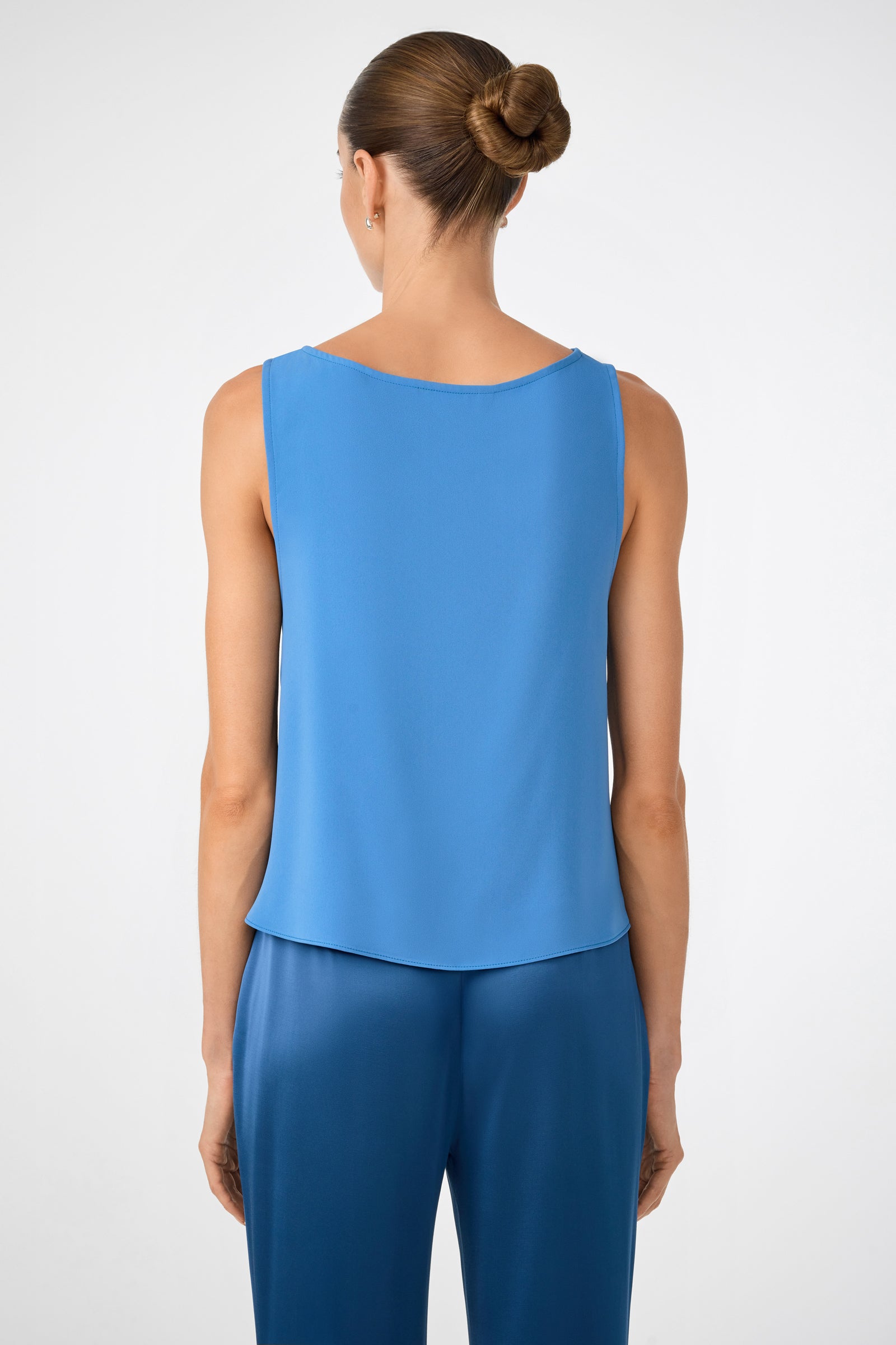 A woman with her hair in a neat bun is seen from behind, wearing the sleeveless blue Kuri Tie-Front Top and matching blue pants, standing against a plain light background.