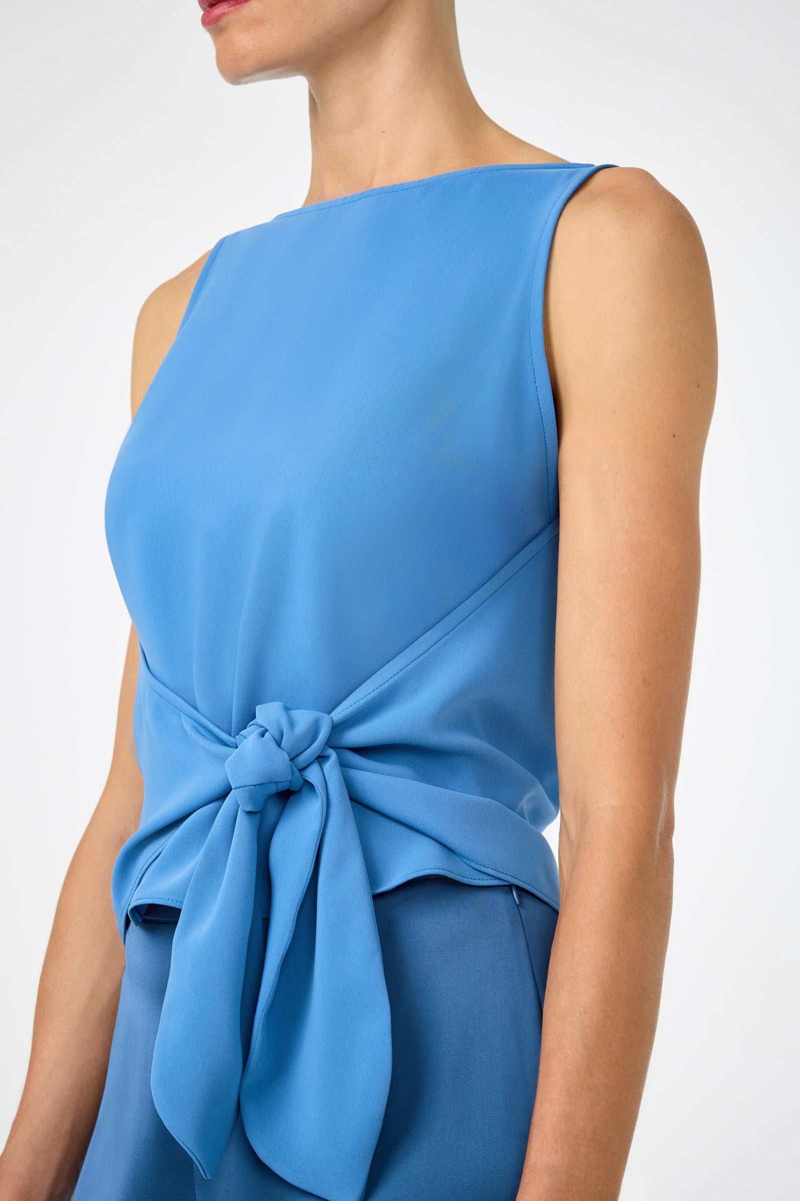 A woman wears the Kuri Tie-Front Top, a sleeveless, boat-neck blue top with a waist tie detail, paired with matching blue pants. Her face is partially out of frame.