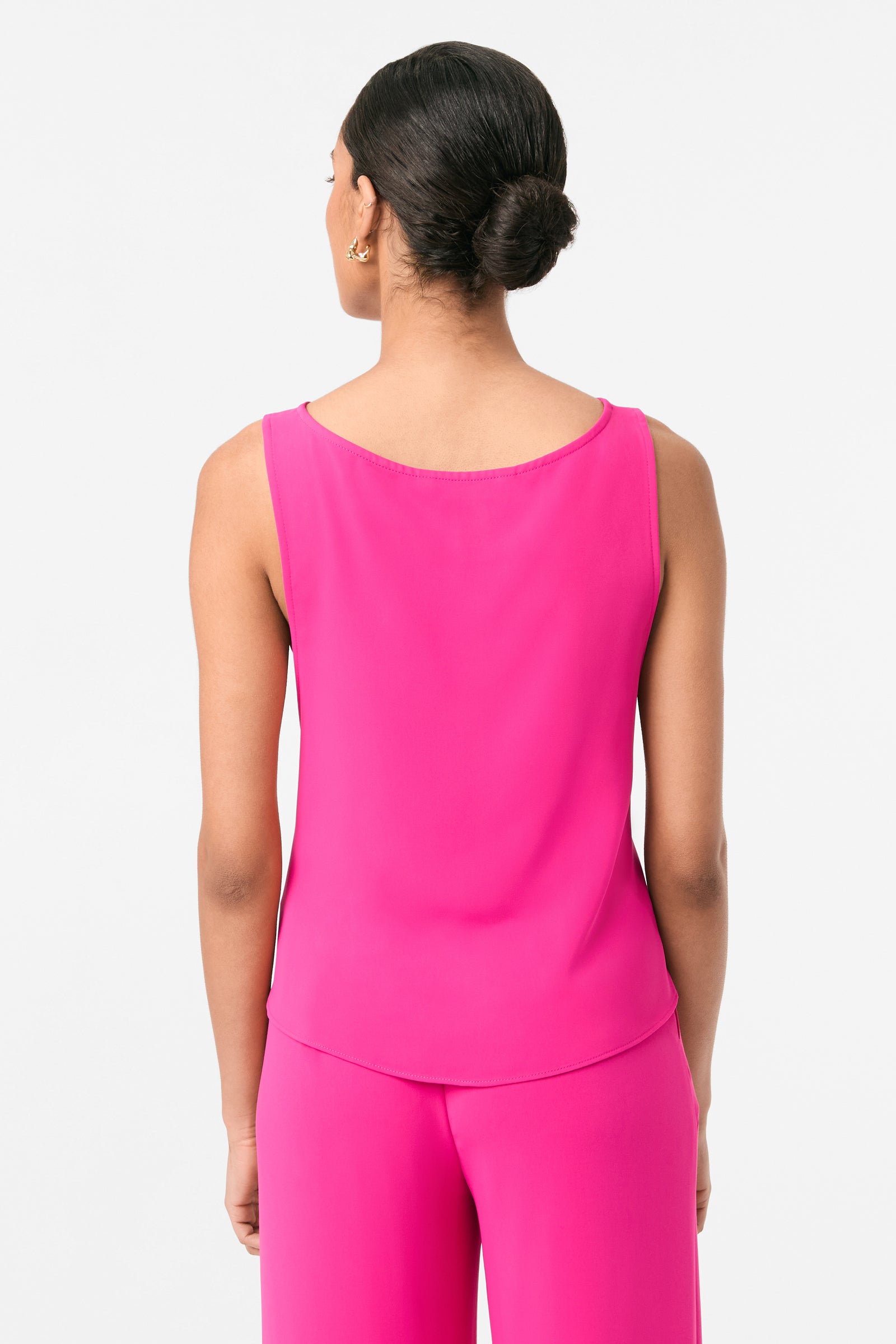A woman with dark hair in a low bun is seen from behind wearing the sleeveless Kuri Tie-Front Top in bright pink, paired with matching pink pants, set against a plain white background.