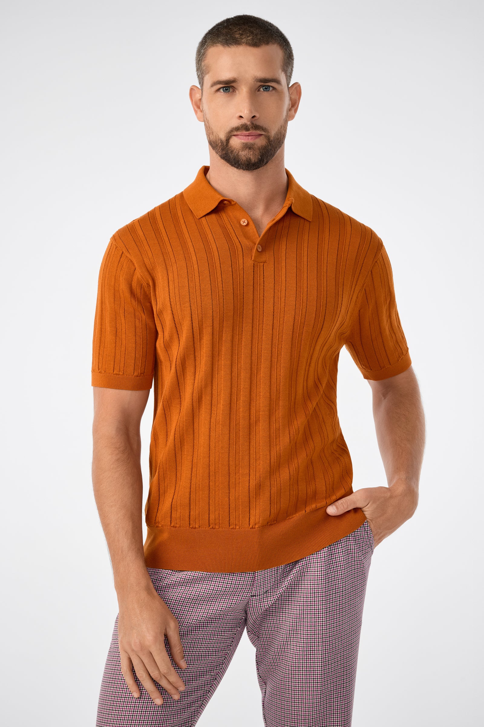 A man with short hair and a beard wears the Kensington Short-Sleeve Polo in Amber and checkered pants, standing against a plain light background with one hand in his pocket.