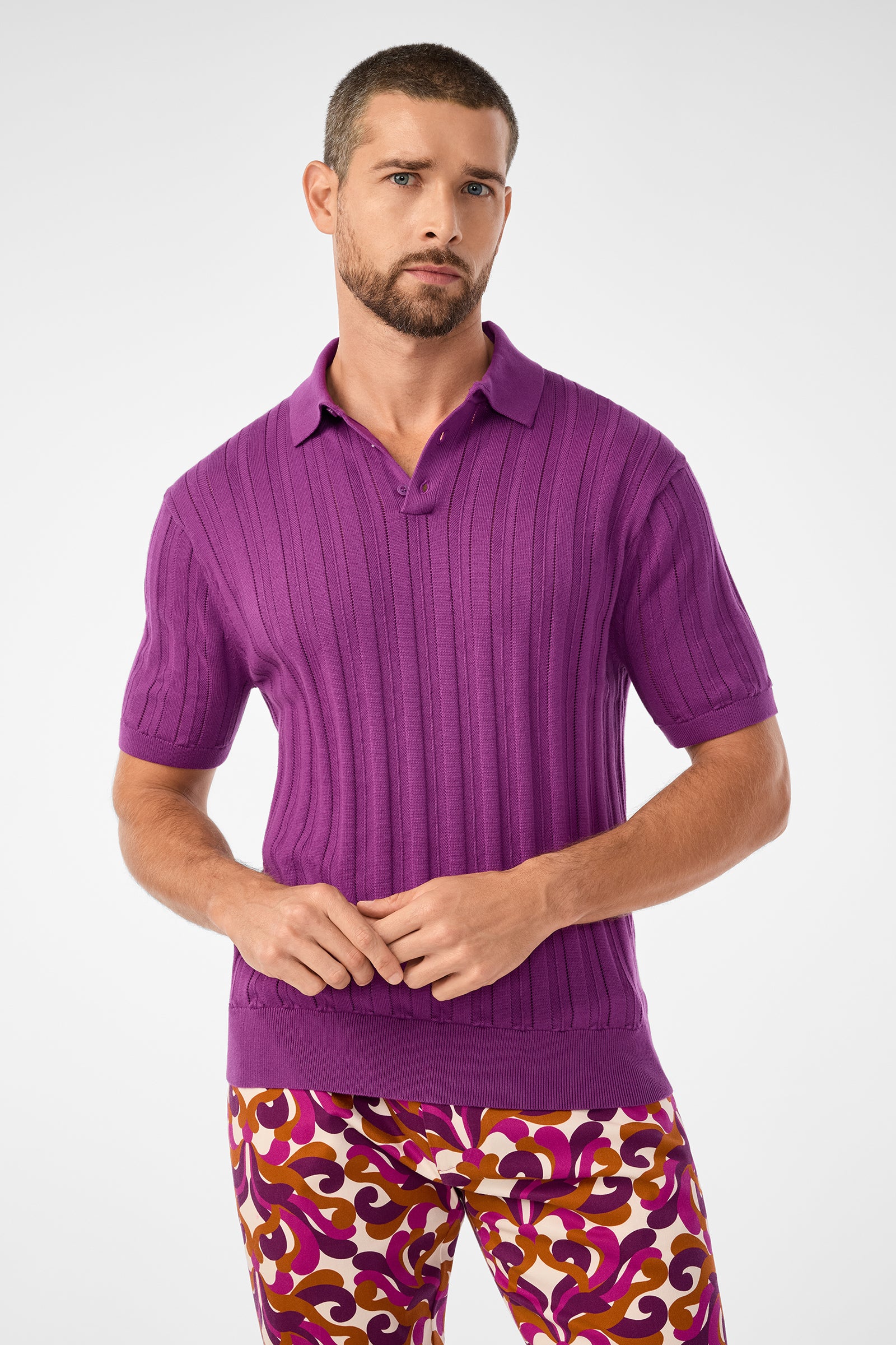 A man with short hair and a trimmed beard wears the Kensington Short-Sleeve Polo in Geranium, paired with vibrant patterned pants in shades of purple, pink, and gold, standing against a plain light background.