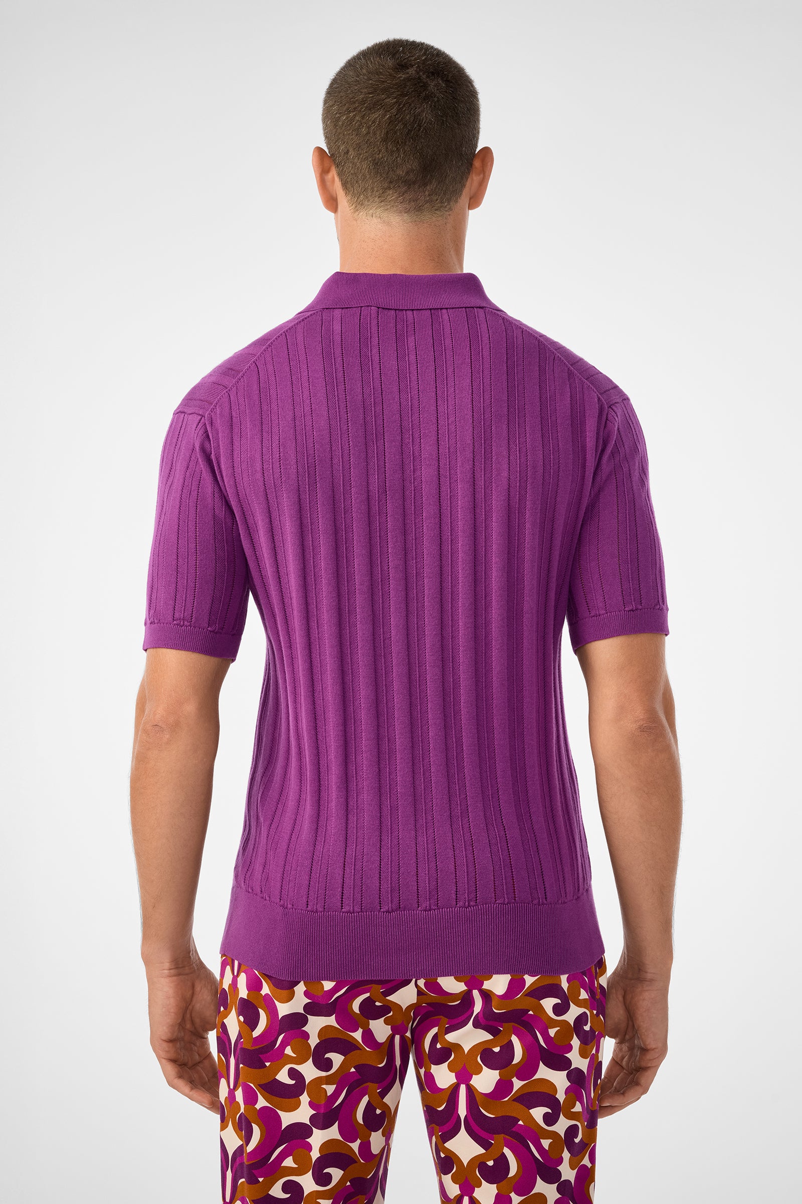 A man stands with his back to the camera, wearing the Kensington Short-Sleeve Polo in Geranium + Amber and colorful swirl-patterned pants. The background is plain and light.